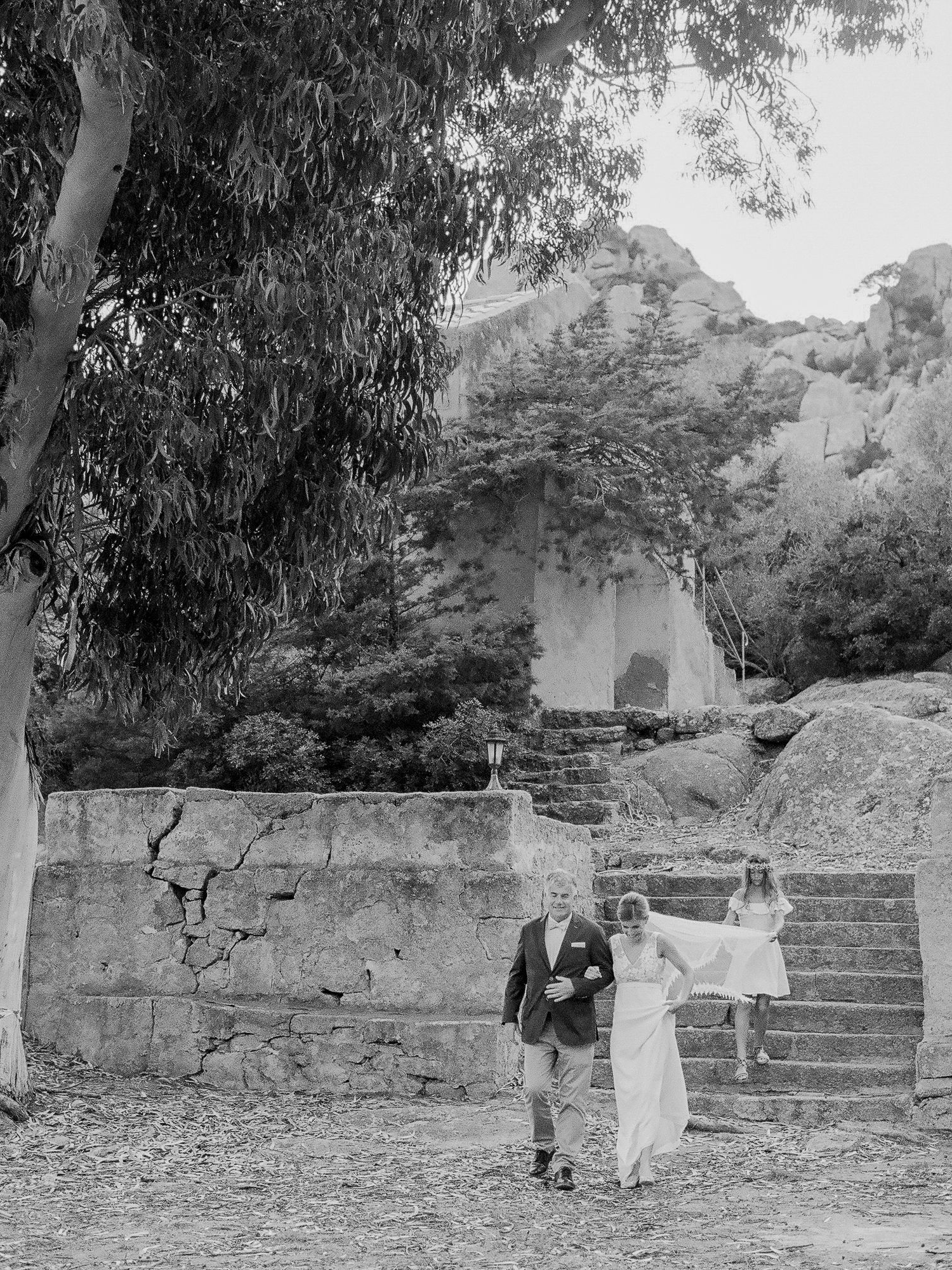 Photo en noir et blanc d'un couple marchant main dans la main à l'extérieur, près de marches en pierre et d'eucalyptus.