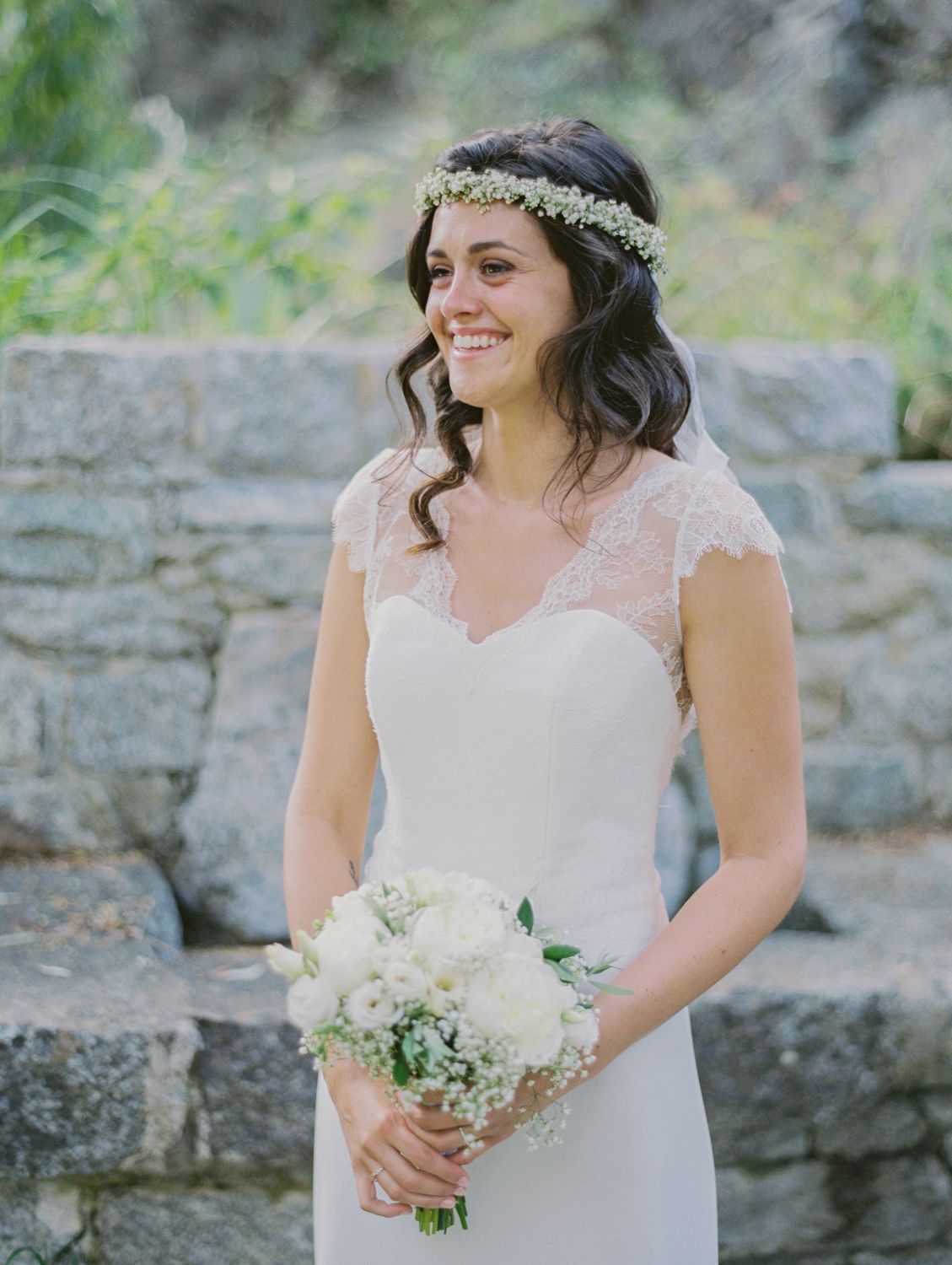 Smiling bride in white lace cap-sleeve gown holds white bouquet, wearing floral crown, standing before stone wall.