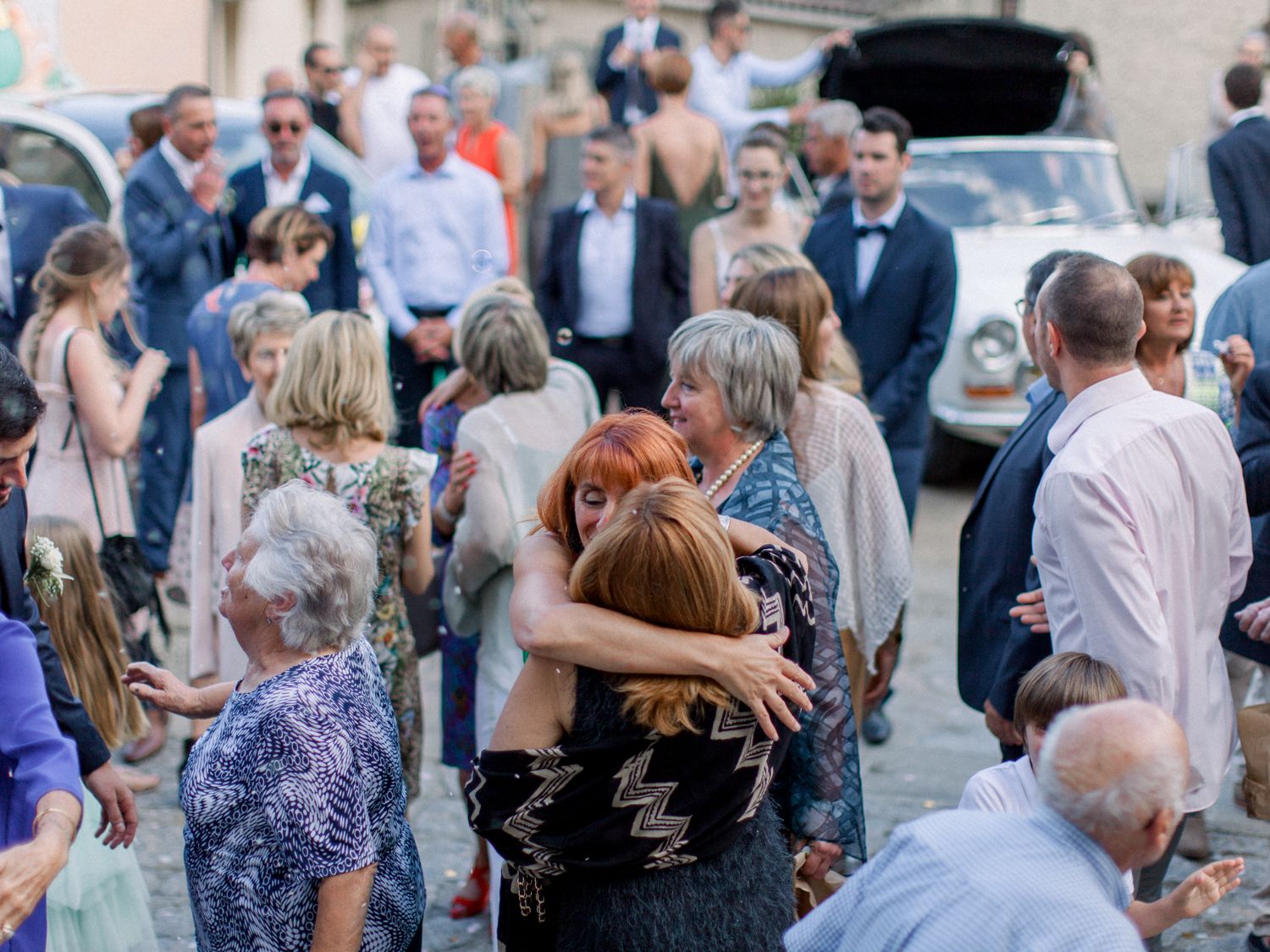 Guests embracing and mingling outdoors at a wedding reception with a vintage car in the background.