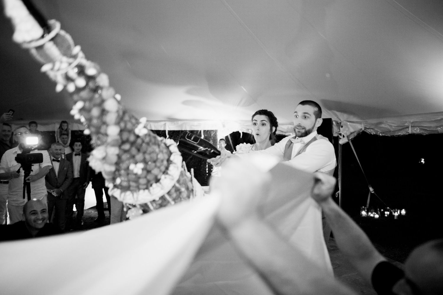 Photo en noir et blanc des mariés coupant leur gâteau de mariage sous une tente lors de leur réception.