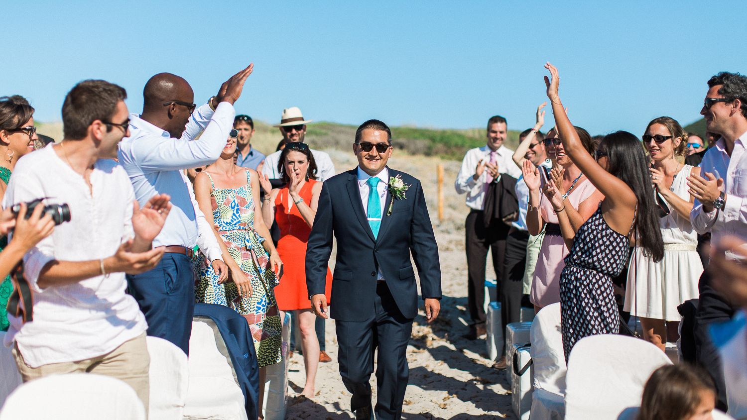 Le marié remonte l'allée de mariage en plein air sur la plage, entouré d'invités en tenue estivale qui l'acclament en Corse