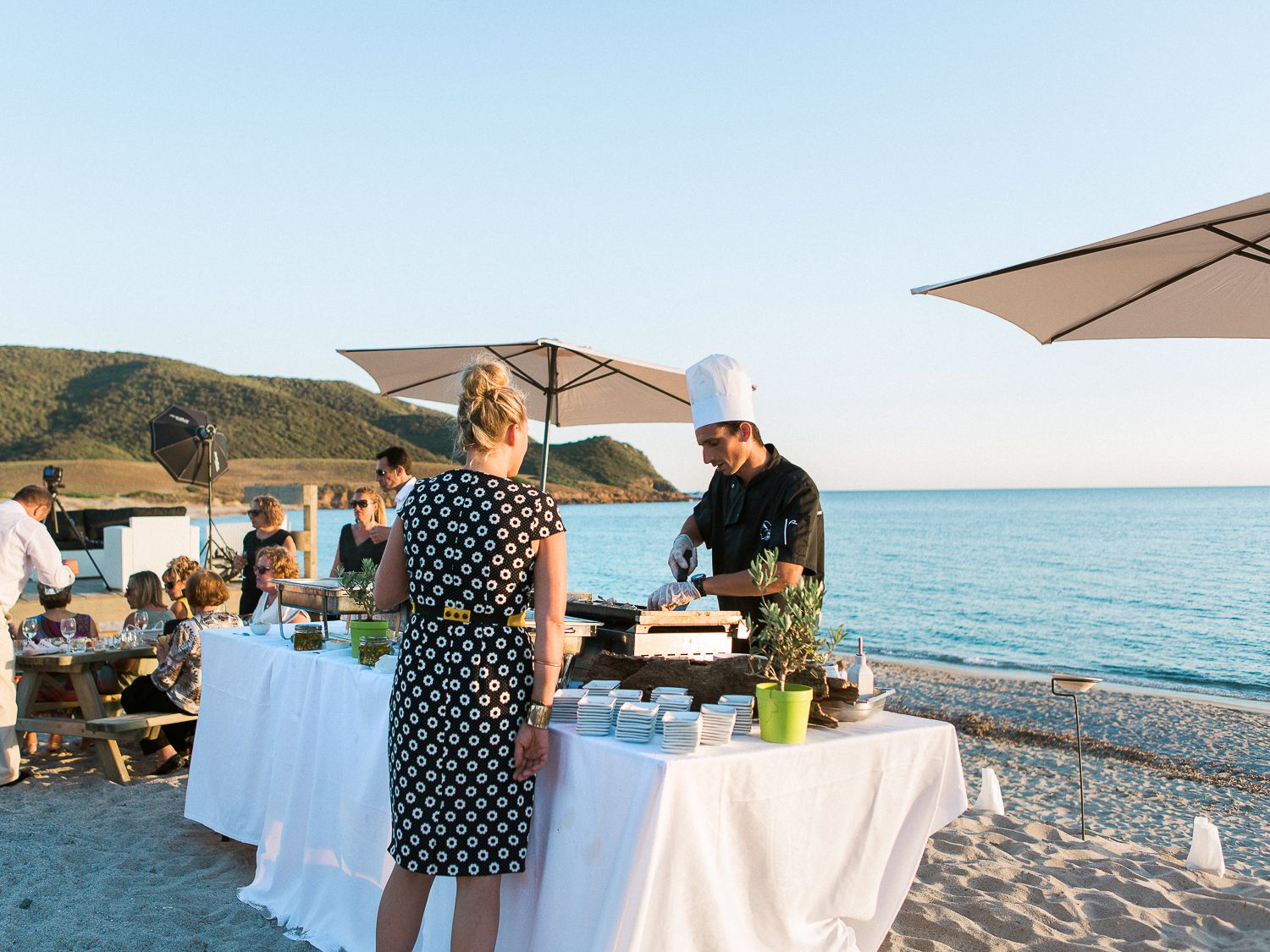 Dîner en plein air sur la plage, avec un chef cuisinant à des tables nappées de blanc, sous des parasols, au coucher du soleil sur la mer.