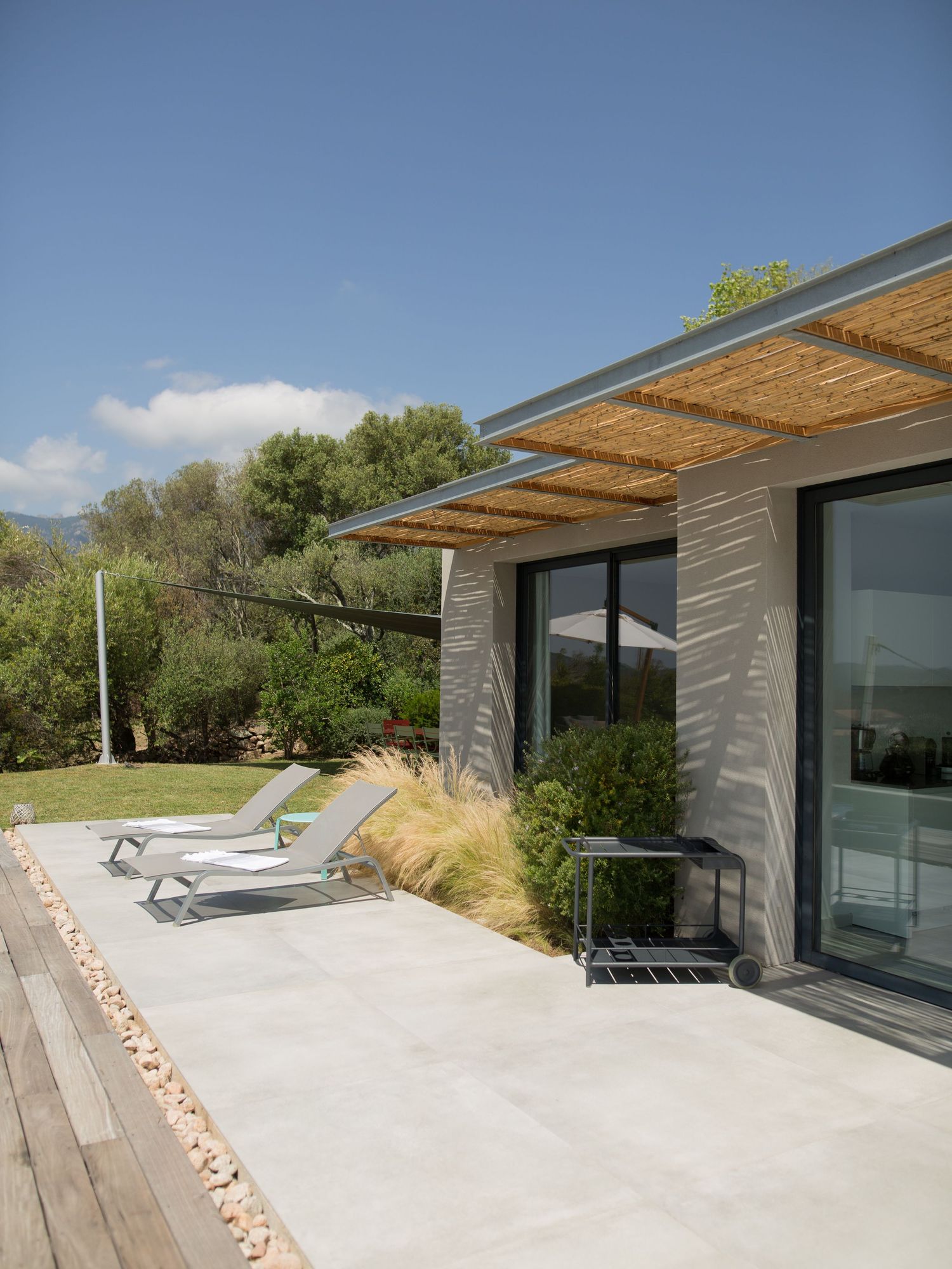 Extérieur de maison contemporaine avec pergola en roseau, chaises longues sur terrasse en pierre et jardin luxuriant.
Muratello.
