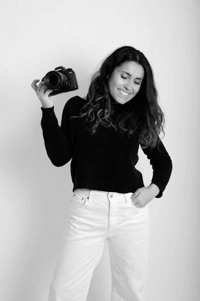 Portrait en noir et blanc d'une femme souriante en col roulé noir et jean blanc tenant un appareil photo.