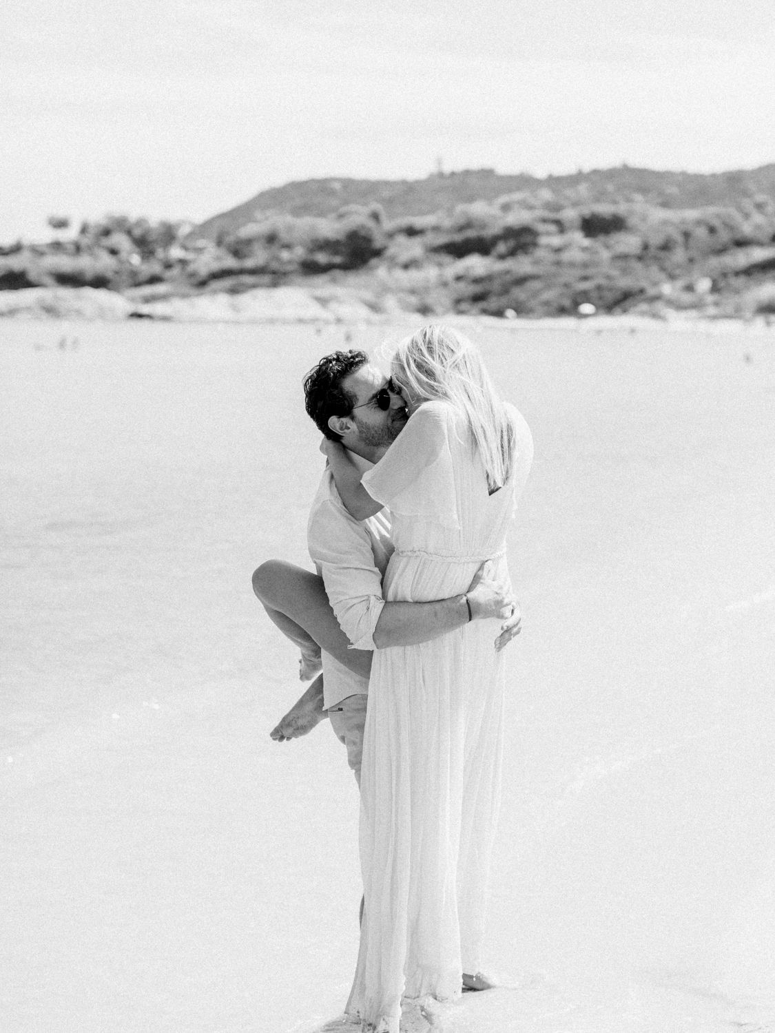 Photo romantique en noir et blanc sur une plage, montrant un marié soulevant sa femme près du rivage à saint Tropez