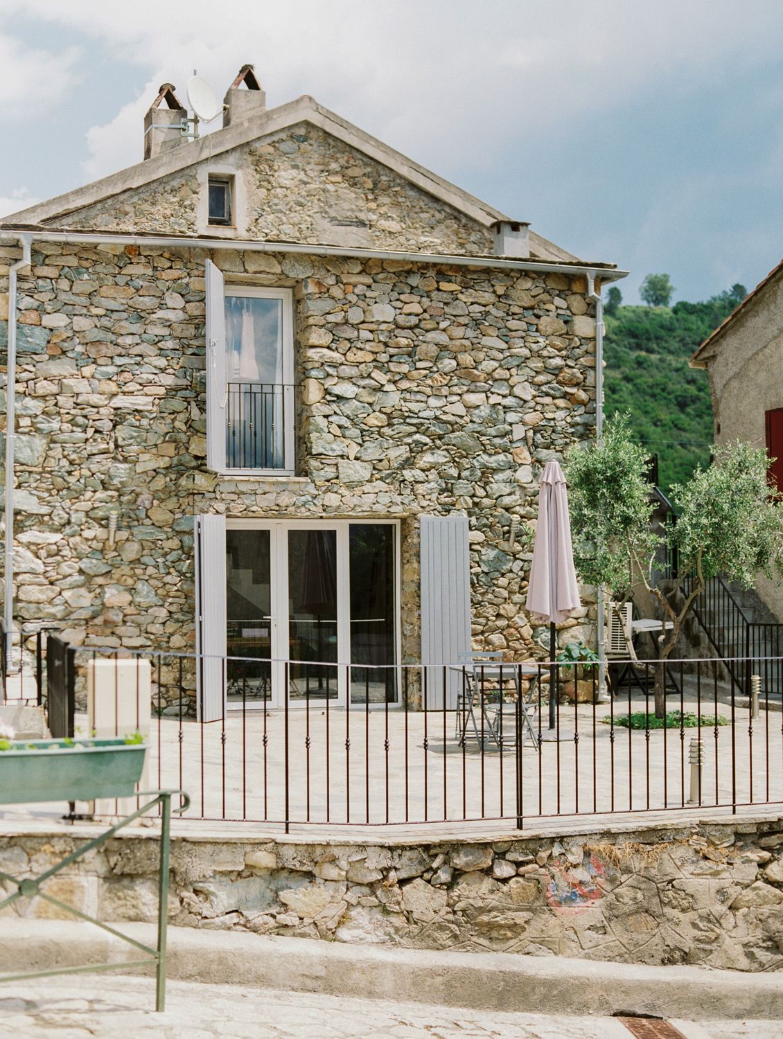 A rustic two-story stone house with French doors, iron fence, and patio furniture nestled in a European village setting.