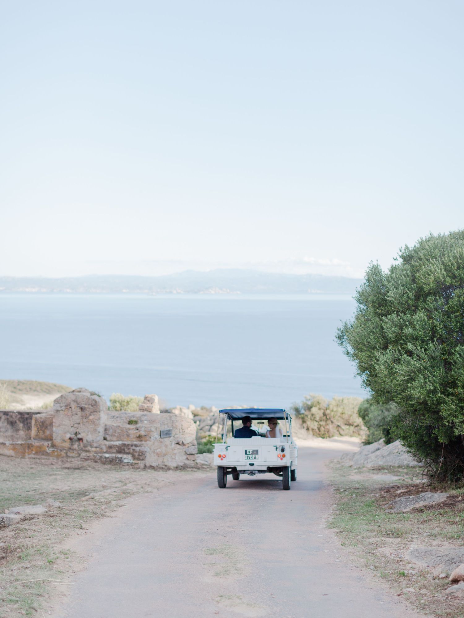 Une voiture d'époque roule sur un chemin côtier bordé de ruines antiques et d'un olivier surplombant une mer bleue calme à Bonifacio