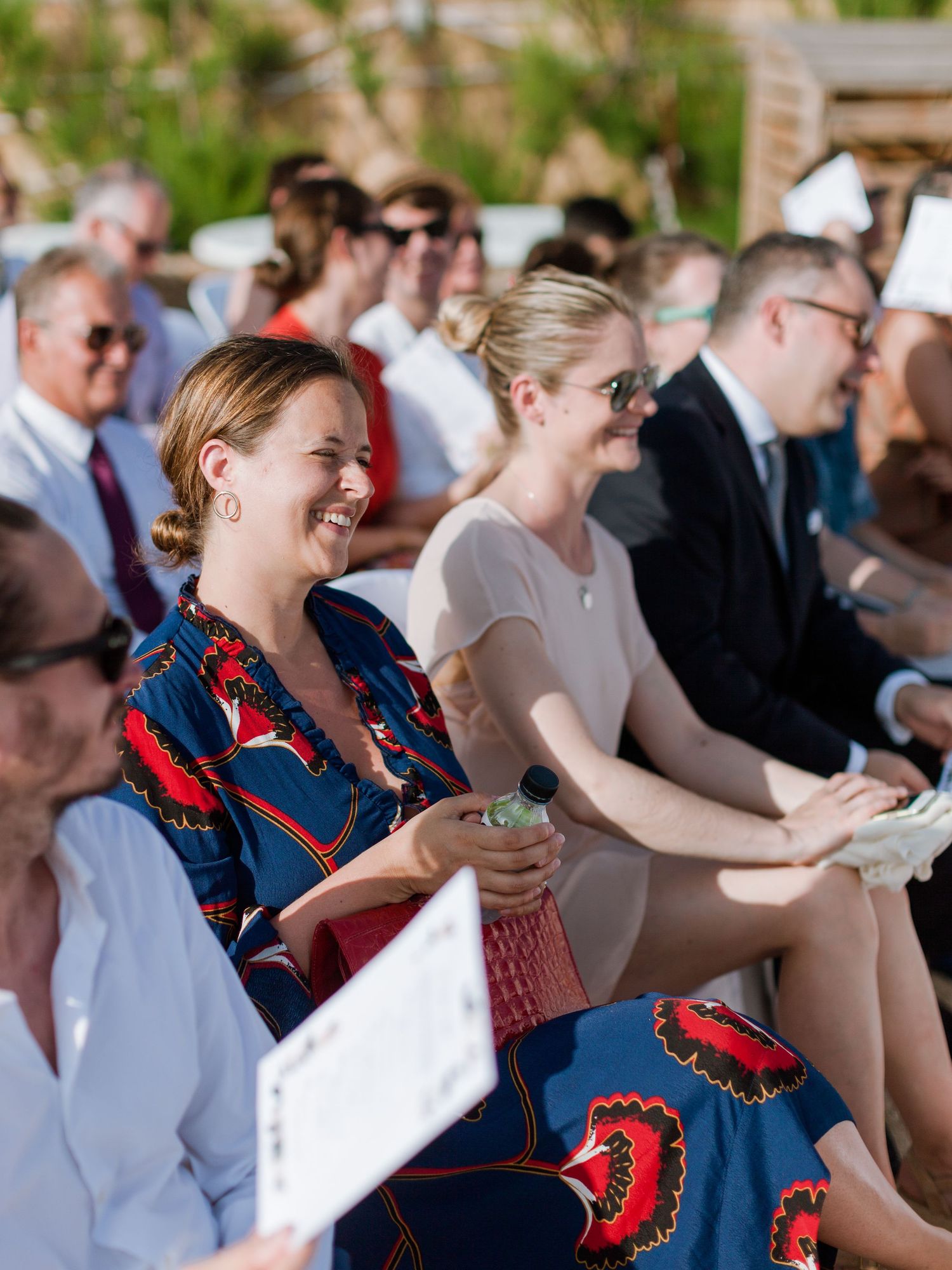 Des invités riant et souriant lors d'une cérémonie de mariage en plein air, vêtus de tenues estivales.