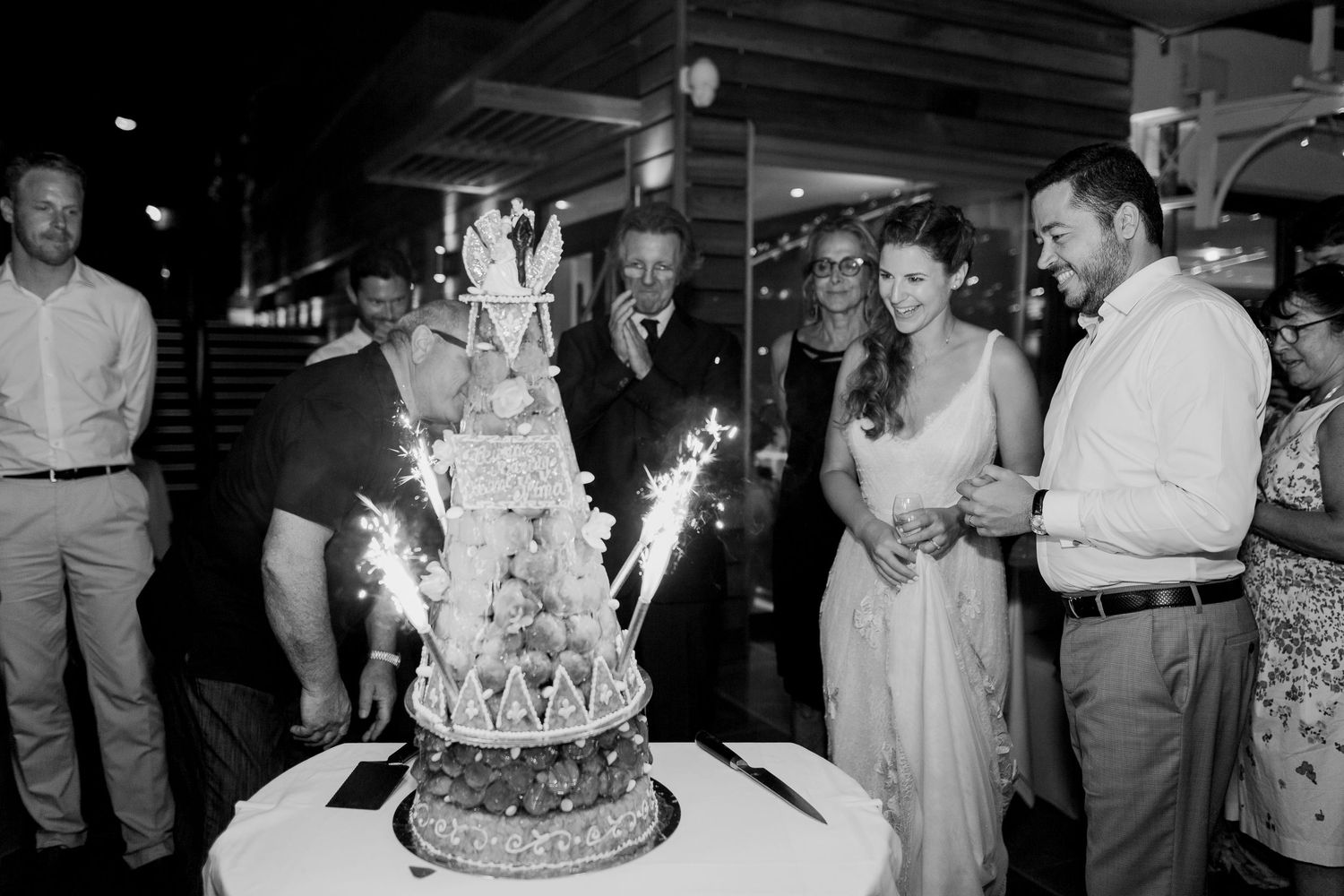 Photo en noir et blanc d'un couple coupant une pièce montée ornée de cierges magiques, entouré d'invités souriants lors d'une réception.