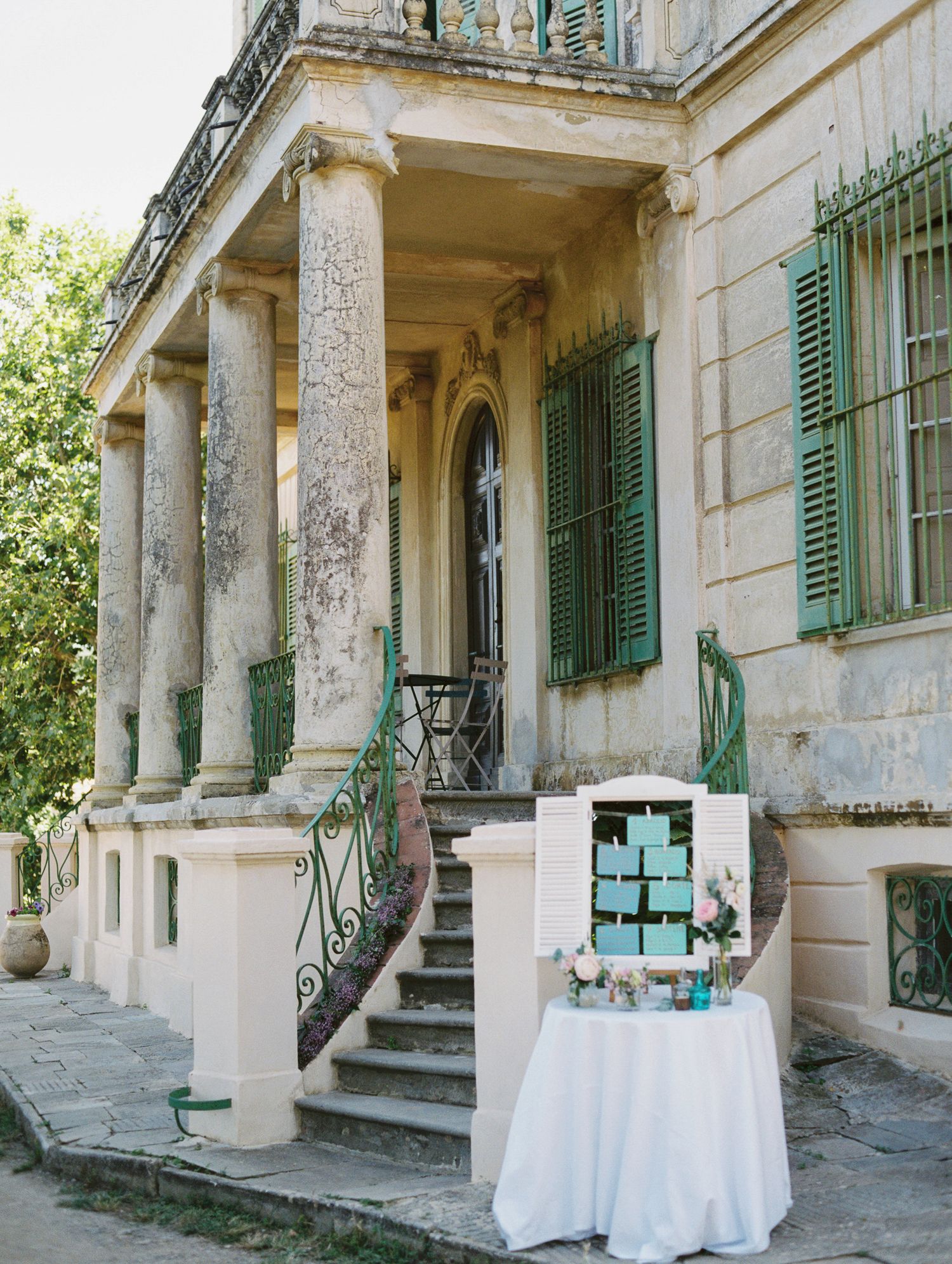 Elegant wedding seating chart display on white table outside a historic villa with green shutters and stone columns.