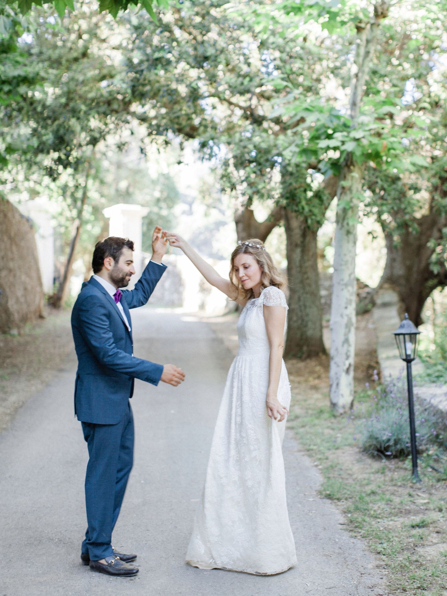 Groom in navy suit twirling bride in white lace gown on tree-lined path at outdoor wedding.