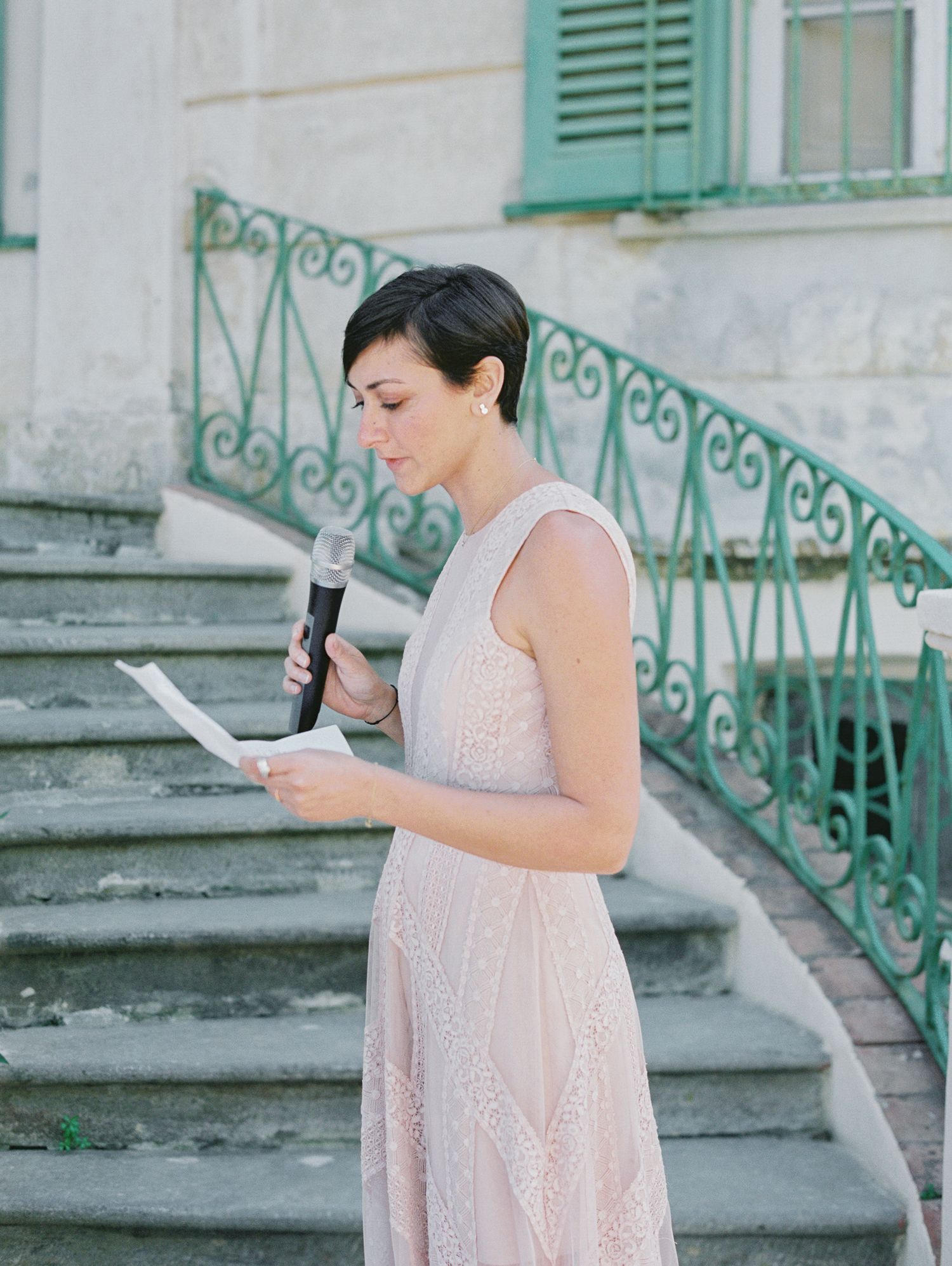 Woman in blush gown reads speech at outdoor wedding ceremony on elegant stone staircase.