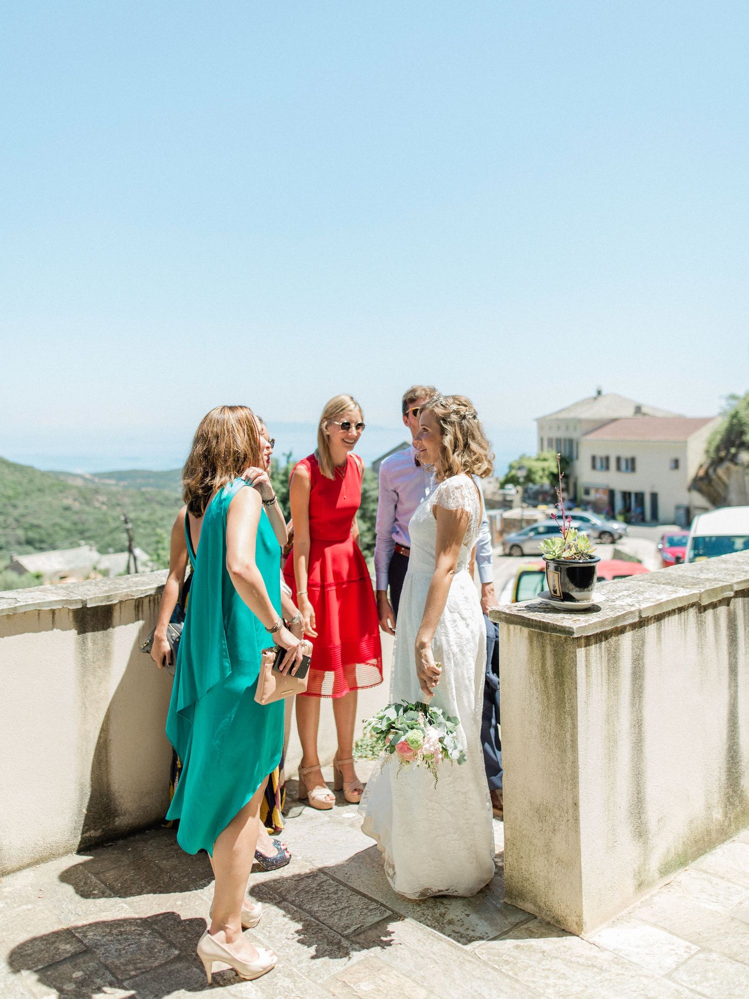 Bride in white gown holds bouquet while chatting with guests in colorful dresses on a sunny outdoor terrace.