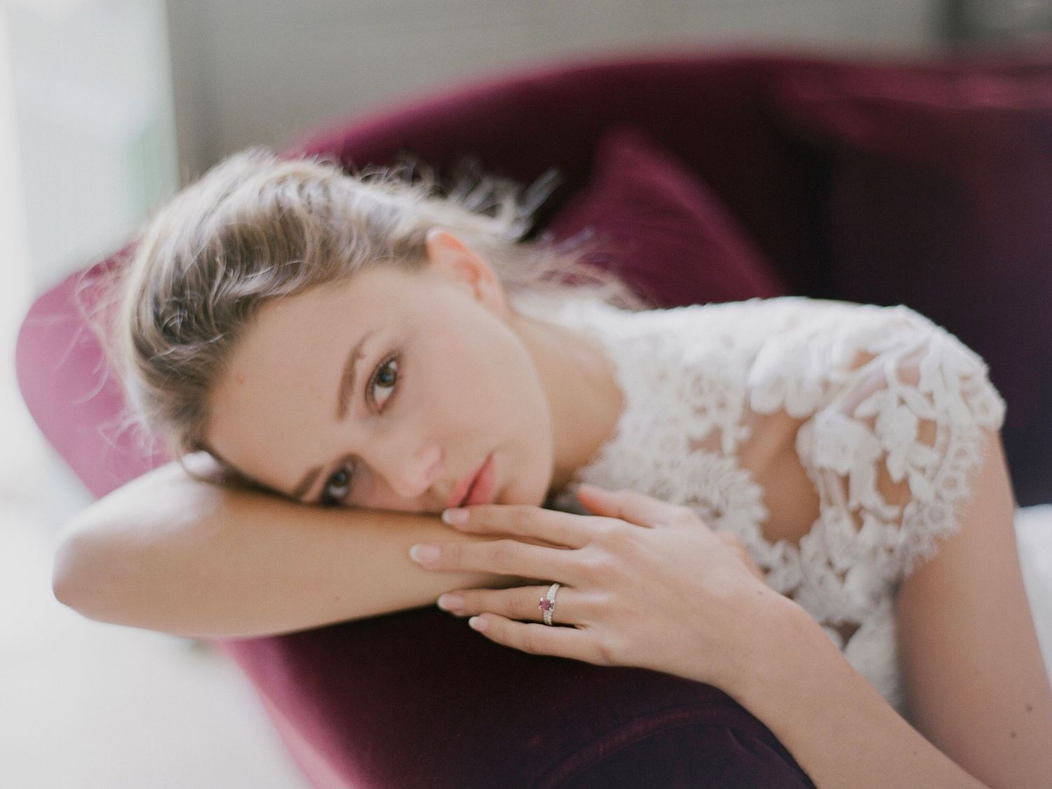 Beautiful bride in lace wedding dress posing on a deep red velvet chair, showcasing her engagement ring.