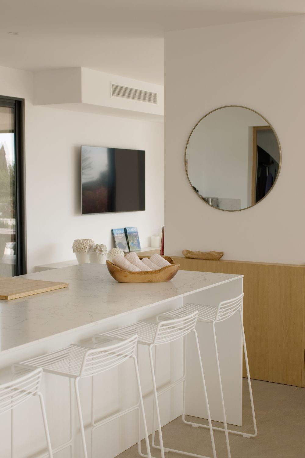 Bar de cuisine blanc contemporain avec tabourets en fil de fer, miroir rond doré, télévision murale et éléments en bois.