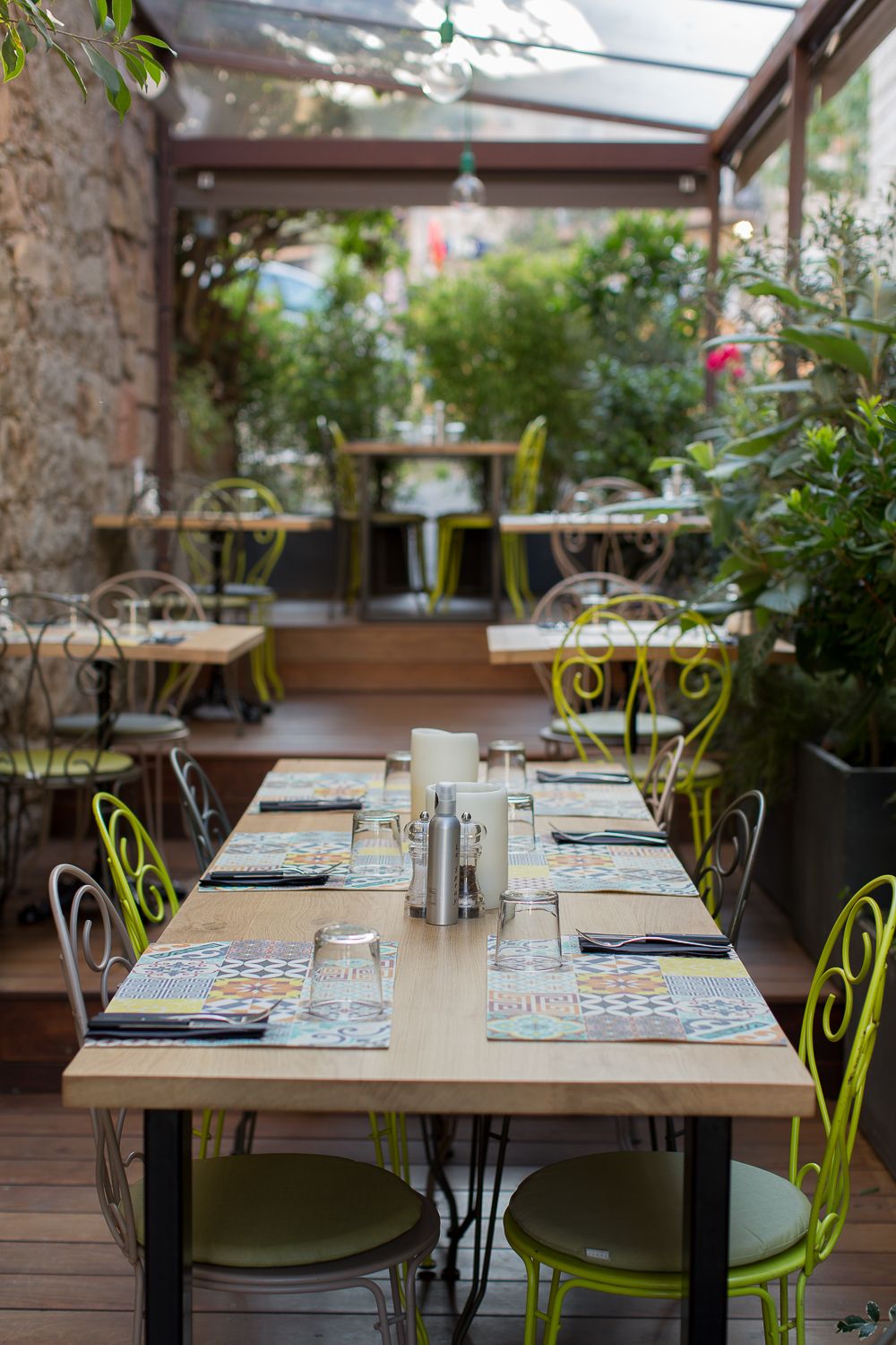 Élégante terrasse de restaurant en plein air avec chaises en métal jaune-vert, tables en bois et végétation luxuriante sous une verrière.