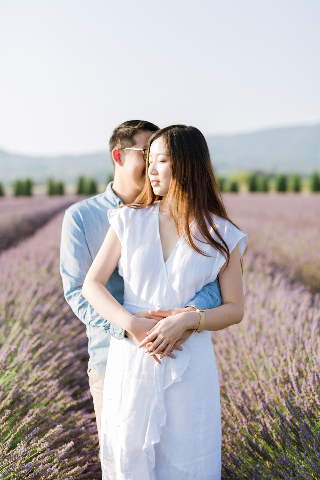 Un couple s'enlaçant dans un champ de lavande, la femme en robe blanche, l'homme en chemise bleue, des fleurs violettes en arrière-plan.
