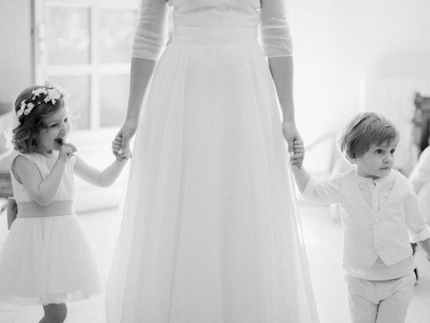 Photo en noir et blanc d'une mariée tenant la main de sa demoiselle d'honneur et de son porteur d'alliances, tous deux vêtus de blanc.