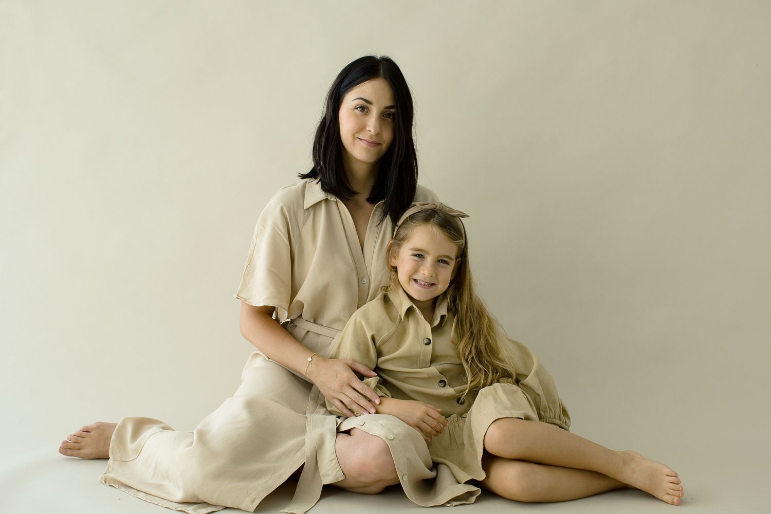 Une mère aux cheveux foncés est assise à côté de sa fille blonde souriante, toutes deux vêtues de tenues assorties en lin beige, en studio.