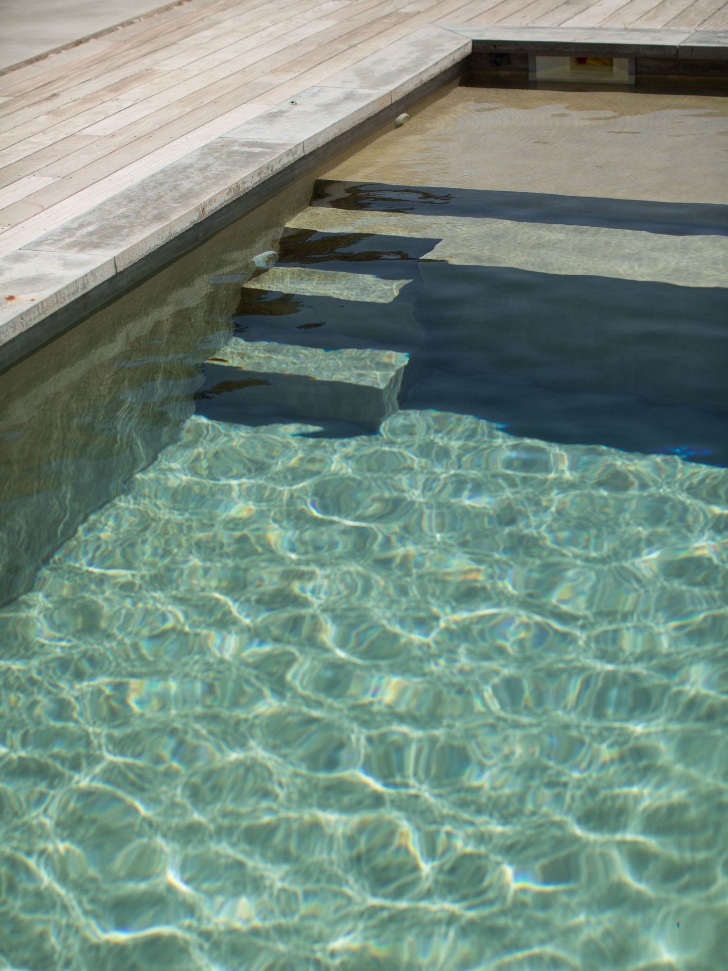 Piscine à l'eau cristalline avec marches immergées et bord de terrasse en bois baigné de soleil. Photoshoot à Porto-vecchio.