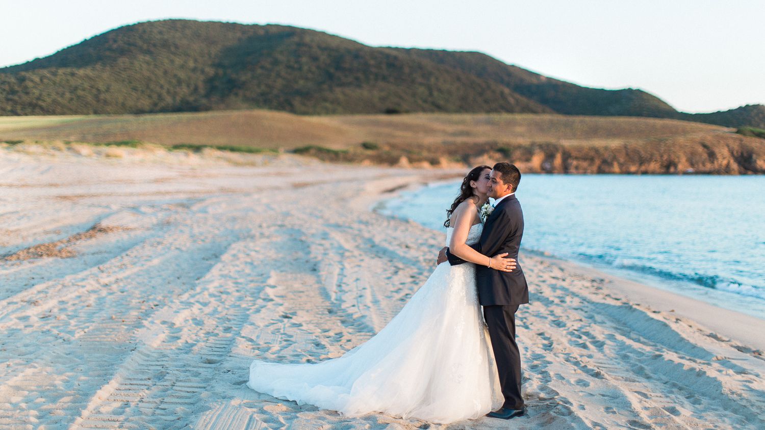 Un couple en tenue de mariage s'enlaçant sur une plage de sable fin, avec une eau turquoise et des collines verdoyantes en arrière-plan.