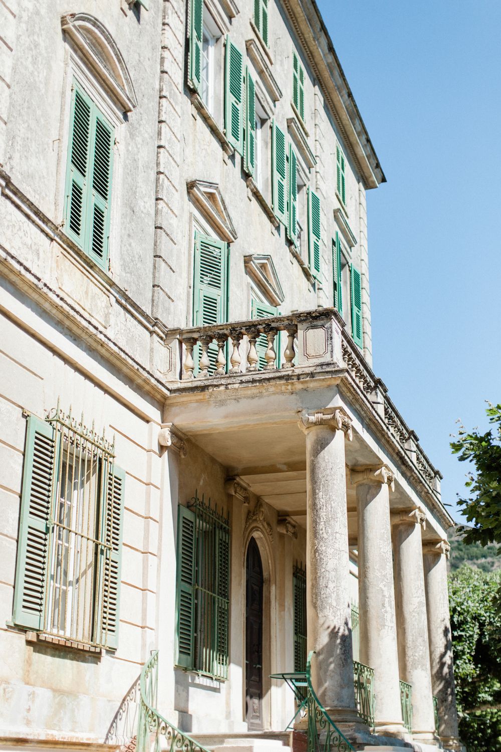 Historic Italian villa with green shutters, ornate balcony, classical columns, and weathered stone facade against blue sky.
