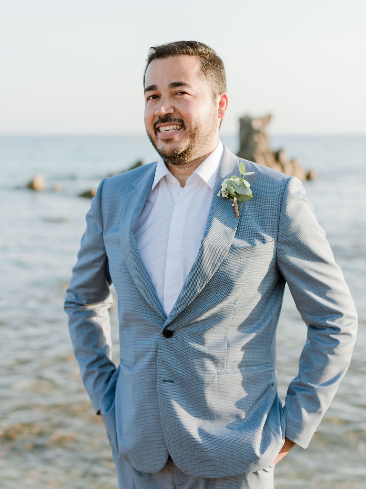 Marié souriant, en costume bleu clair avec une boutonnière, debout près des rochers en bord de mer lors d'un mariage sur la plage.