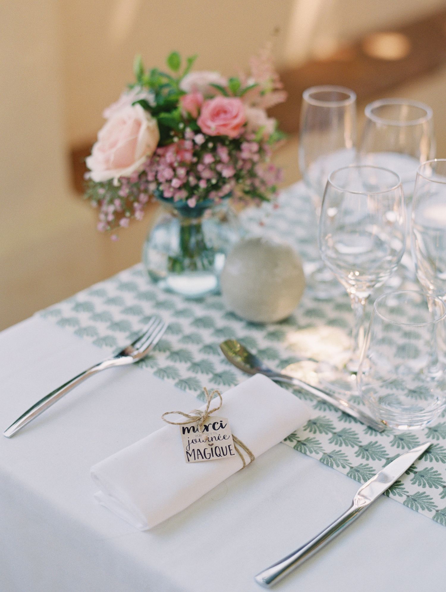 Elegant wedding table setting with pink roses, crystal glasses, white napkin with gift tag, and green patterned runner.