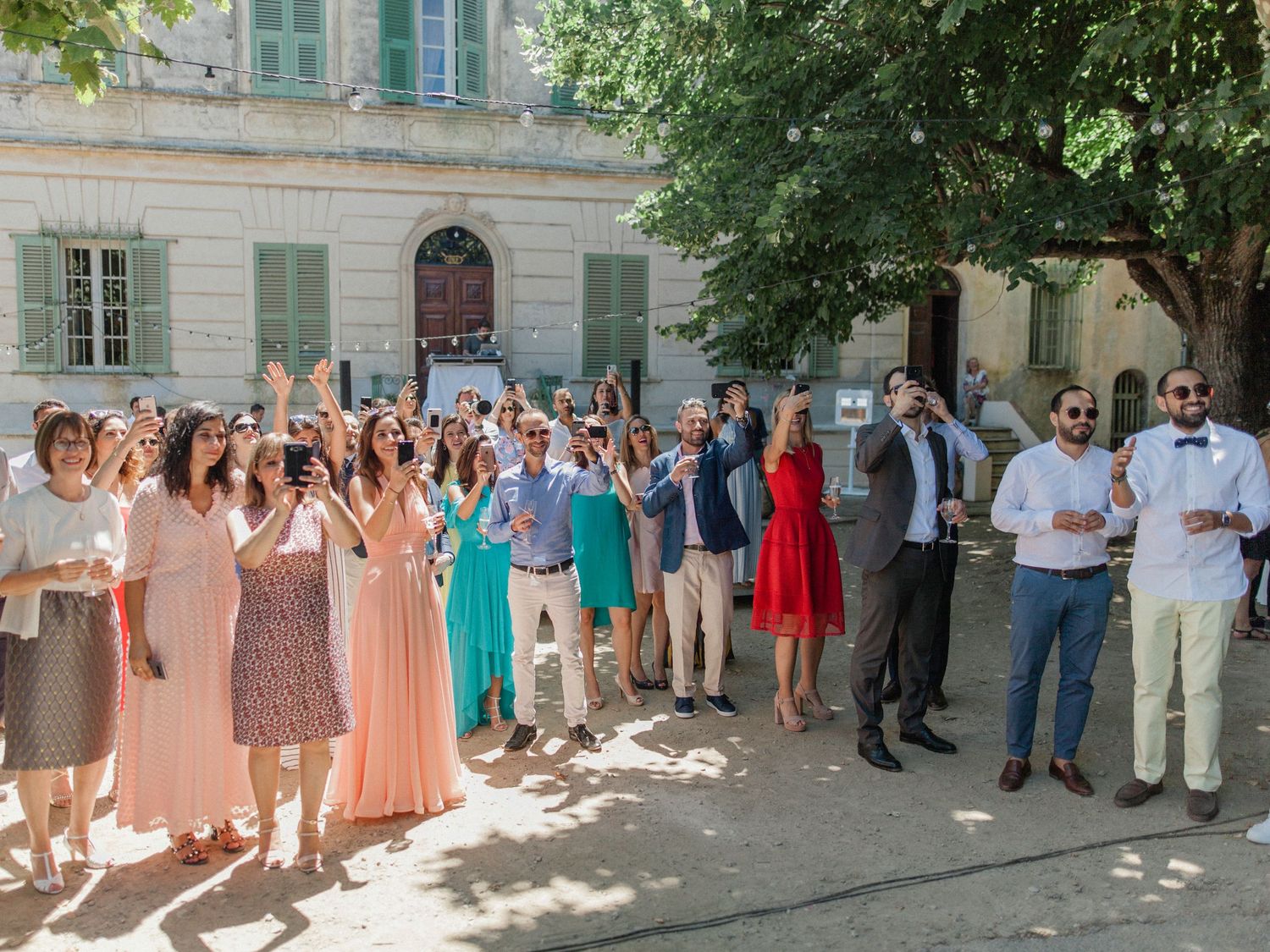 Large group of wedding guests celebrating outdoors in front of a historic building on a sunny day.