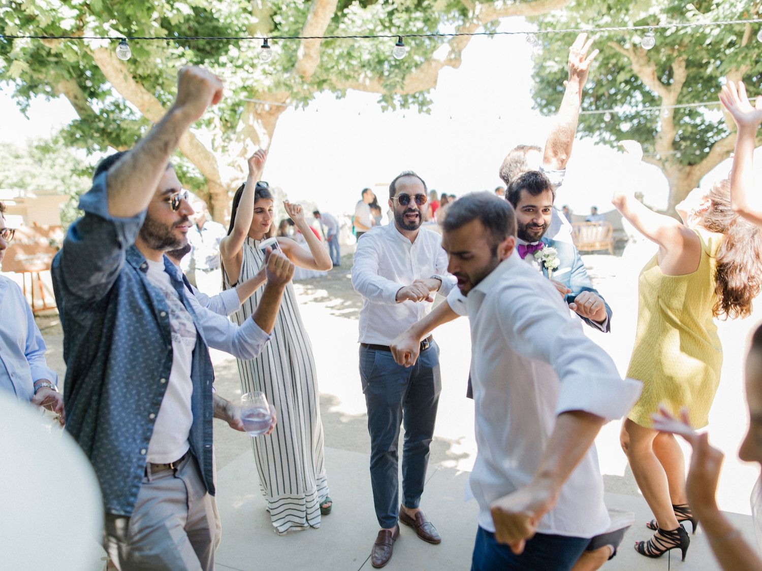 Guests dancing joyfully outdoors at a summer wedding celebration under trees with string lights.