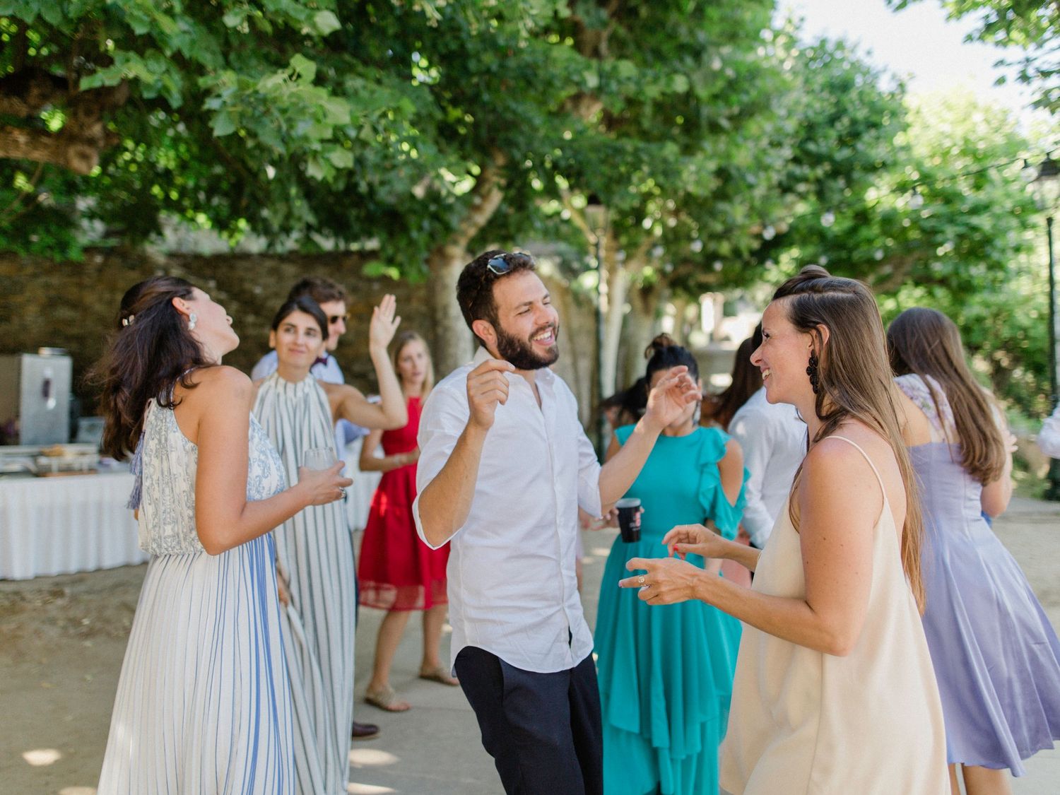 Guests dancing and laughing outdoors at a summer wedding reception under green trees.