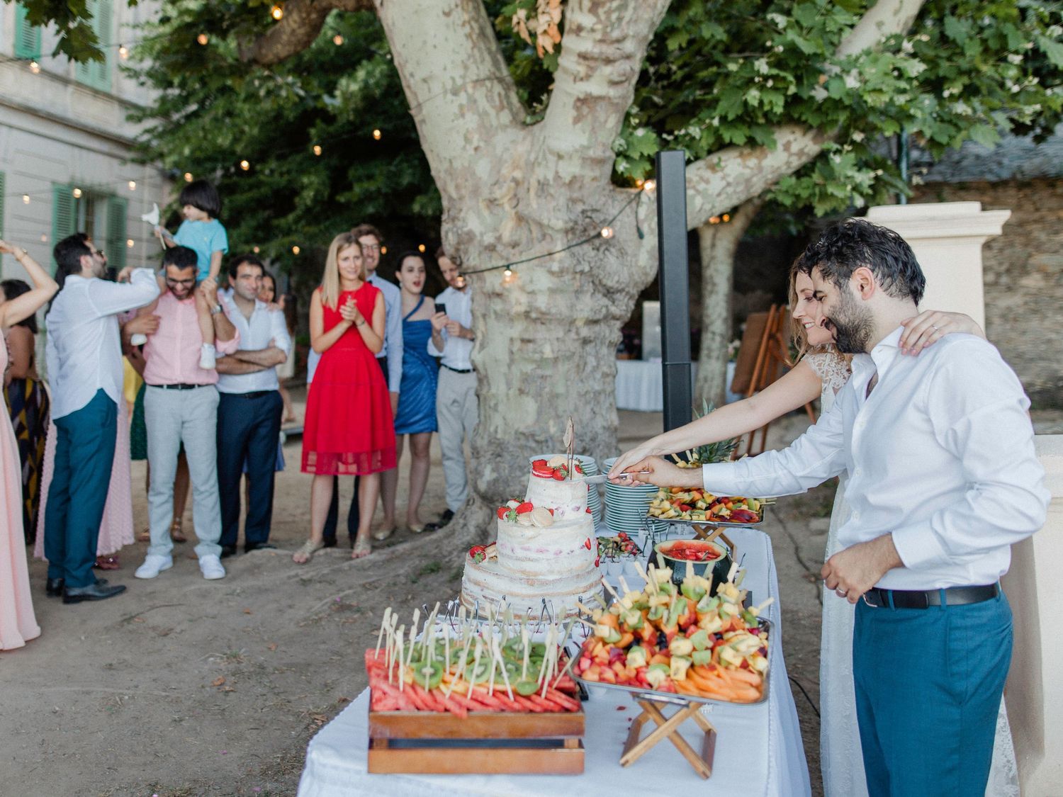 Couple cutting wedding cake outdoors at elegant garden reception with guests celebrating under string lights.