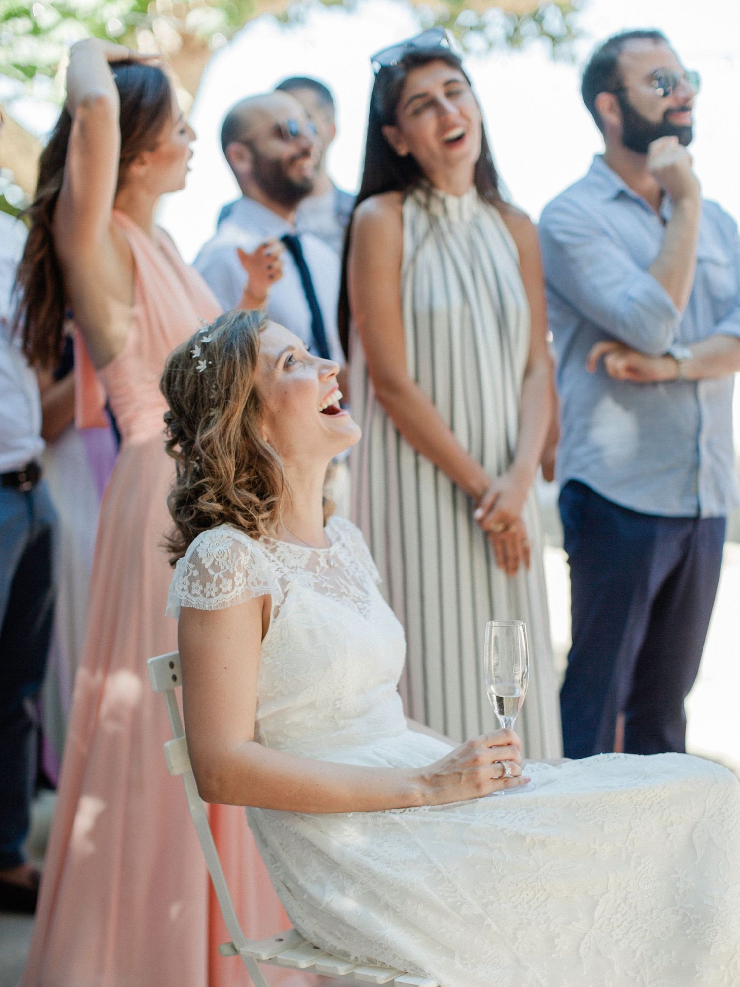 Bride in white lace dress laughing joyfully at wedding reception, holding champagne glass, guests in background.