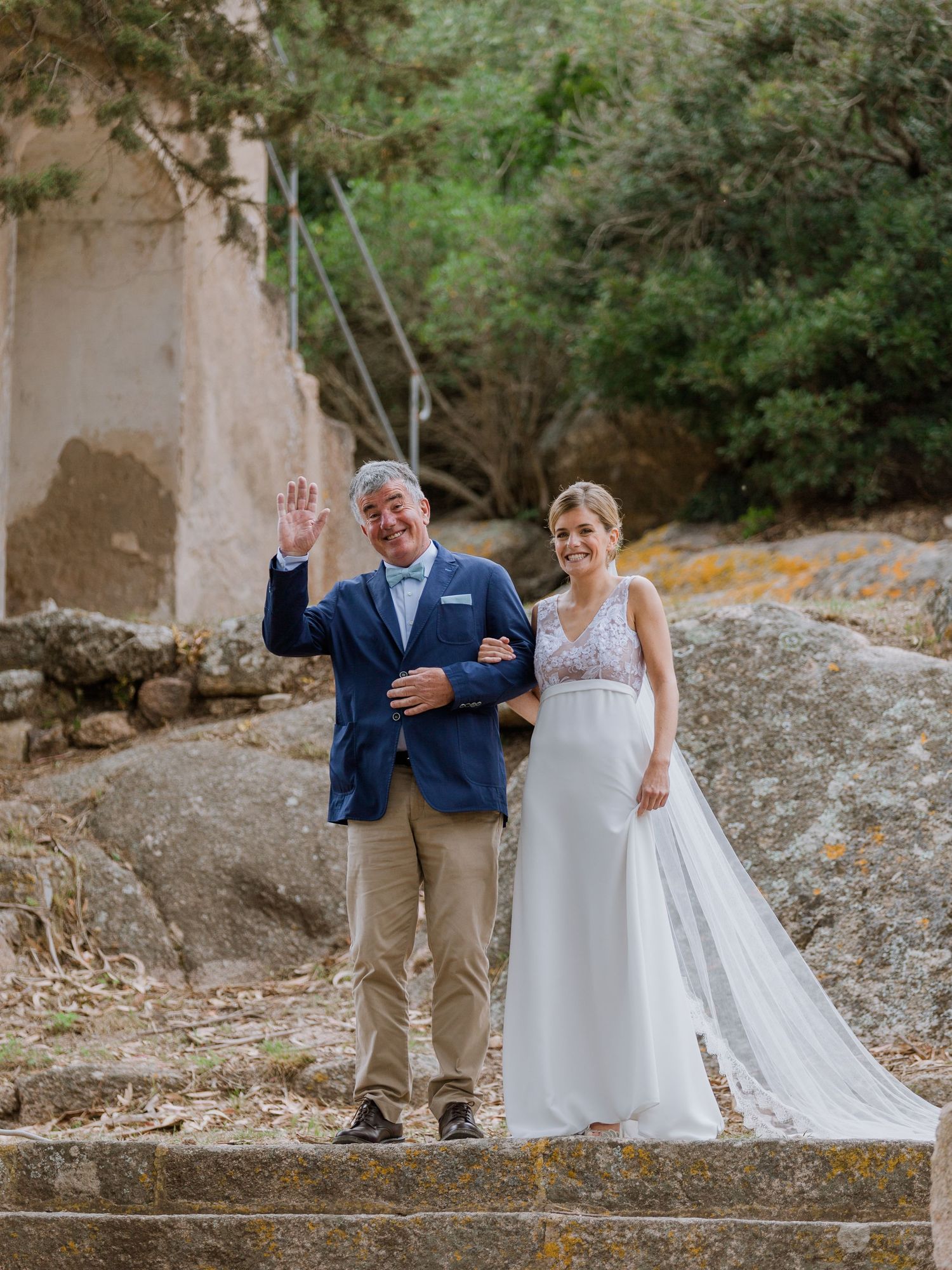 Un couple souriant en tenue de mariage pose sur des marches en pierre à l'extérieur, le marié en blazer bleu faisant un signe de la main
