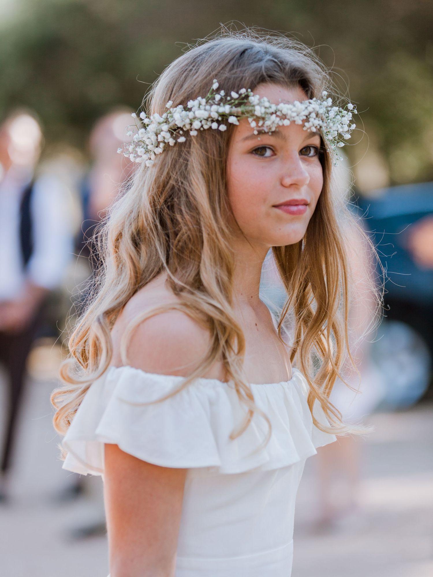 Jeune fille en robe blanche à épaules dénudées portant une couronne de gypsophile lors d'une cérémonie de mariage en plein air.