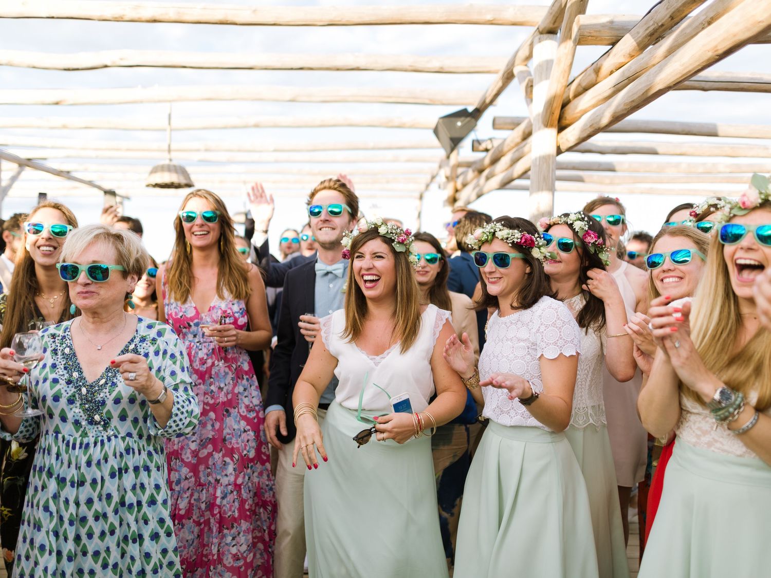 Des invités joyeux, portant des lunettes de soleil colorées à verres miroirs, célèbrent un mariage en plein air sous une pergola rustique