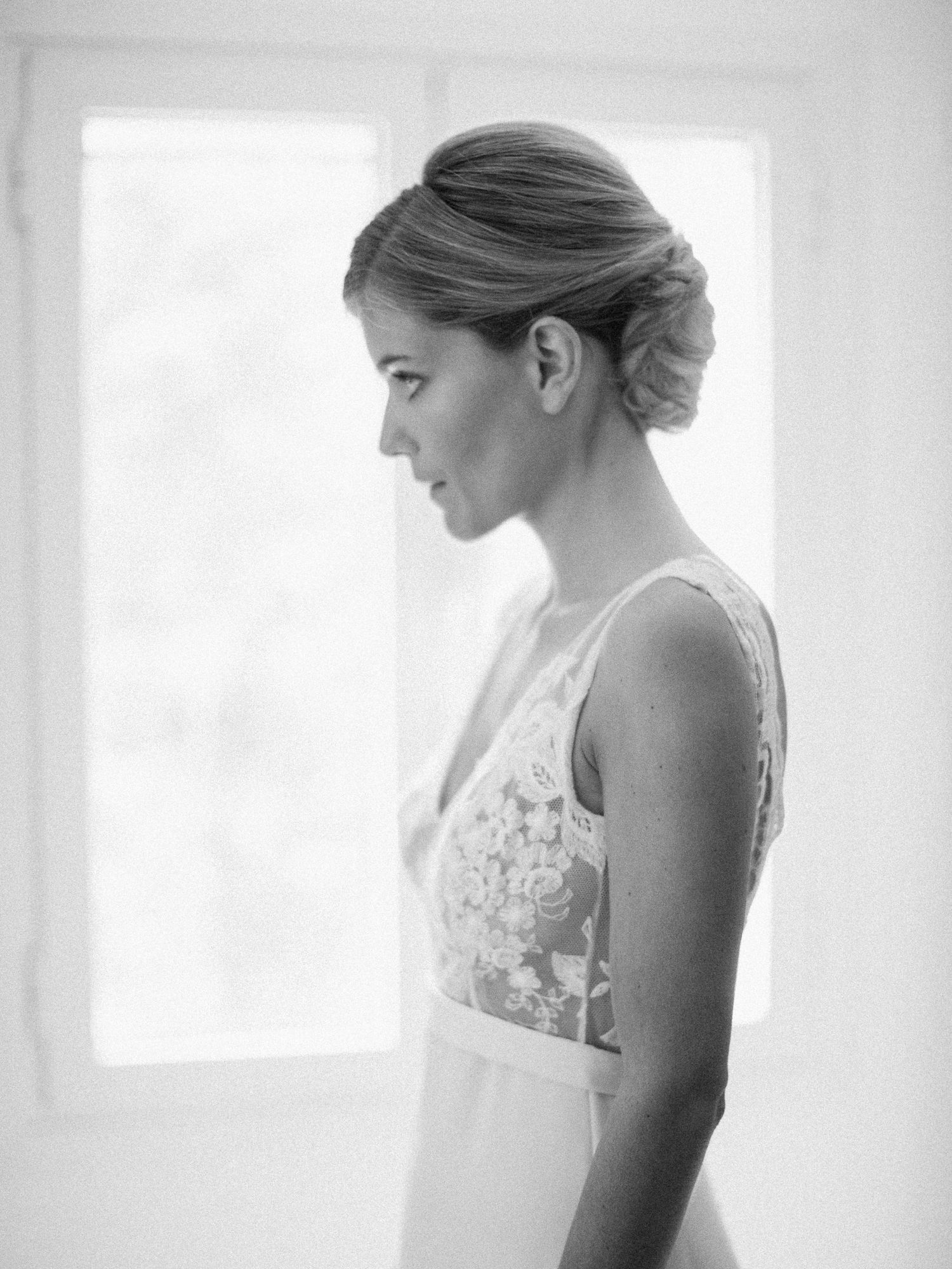 Portrait de profil en noir et blanc d'une mariée en robe de mariée en dentelle, coiffée d'un élégant chignon, près d'une fenêtre.