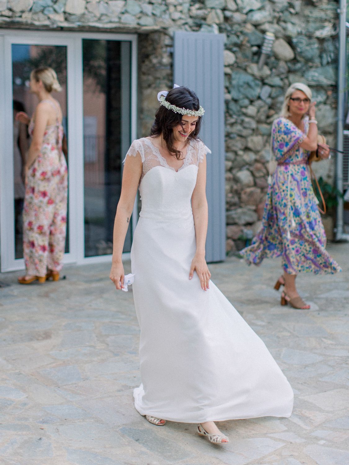 Bride in white lace cap-sleeve wedding gown and floral crown smiles outdoors at stone villa wedding venue.