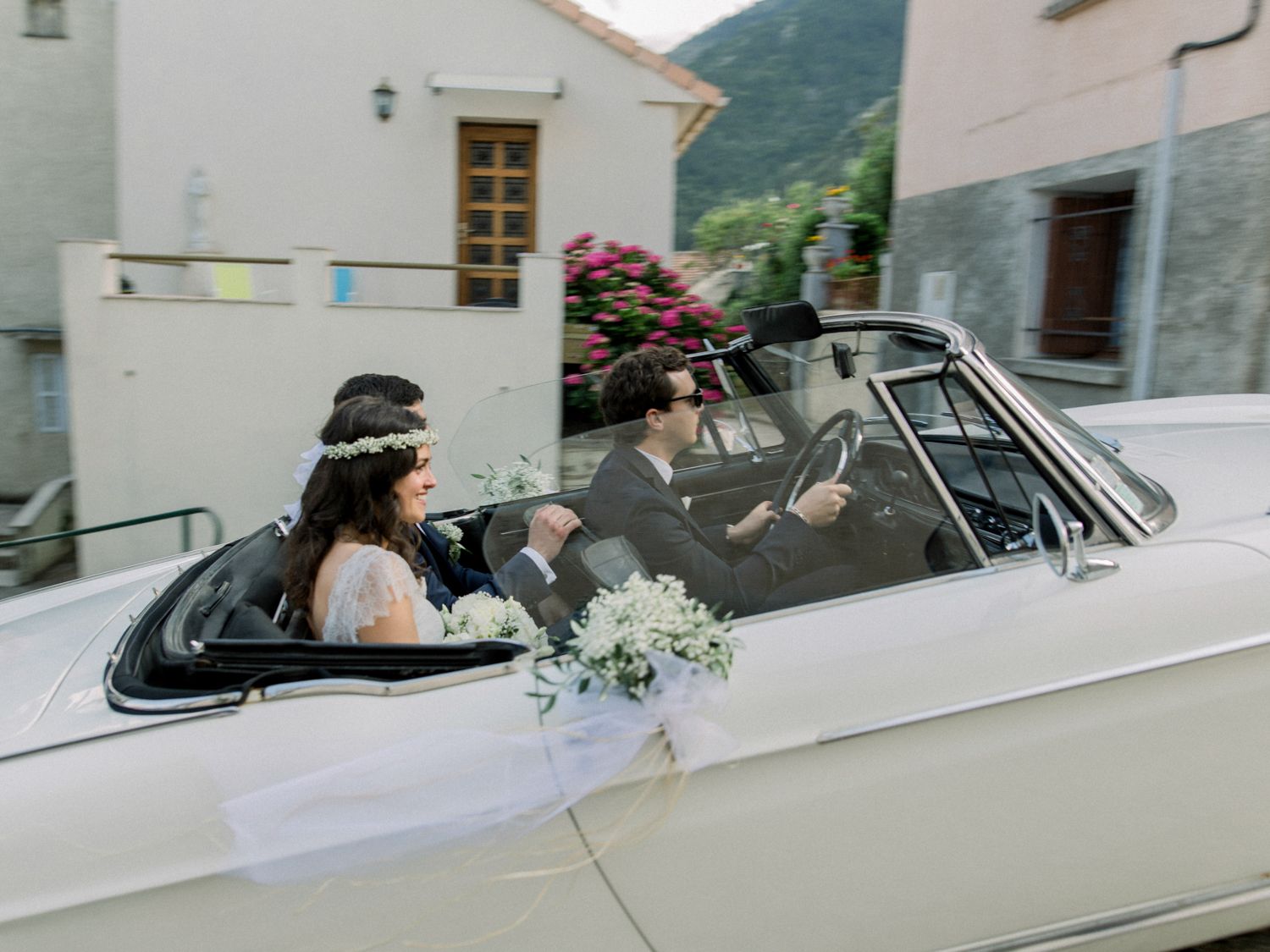 Bride and groom riding in a decorated vintage white convertible car with floral bouquet on display.