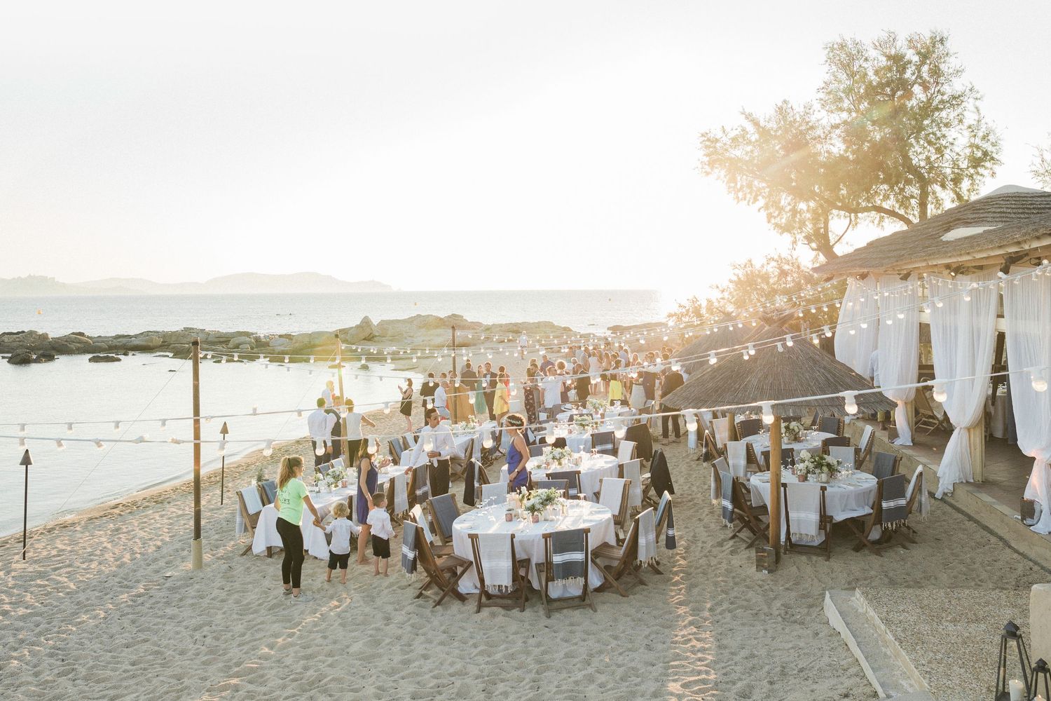 Elegant beach wedding reception at sunset with round tables, white drapes, string lights, and mountain views.