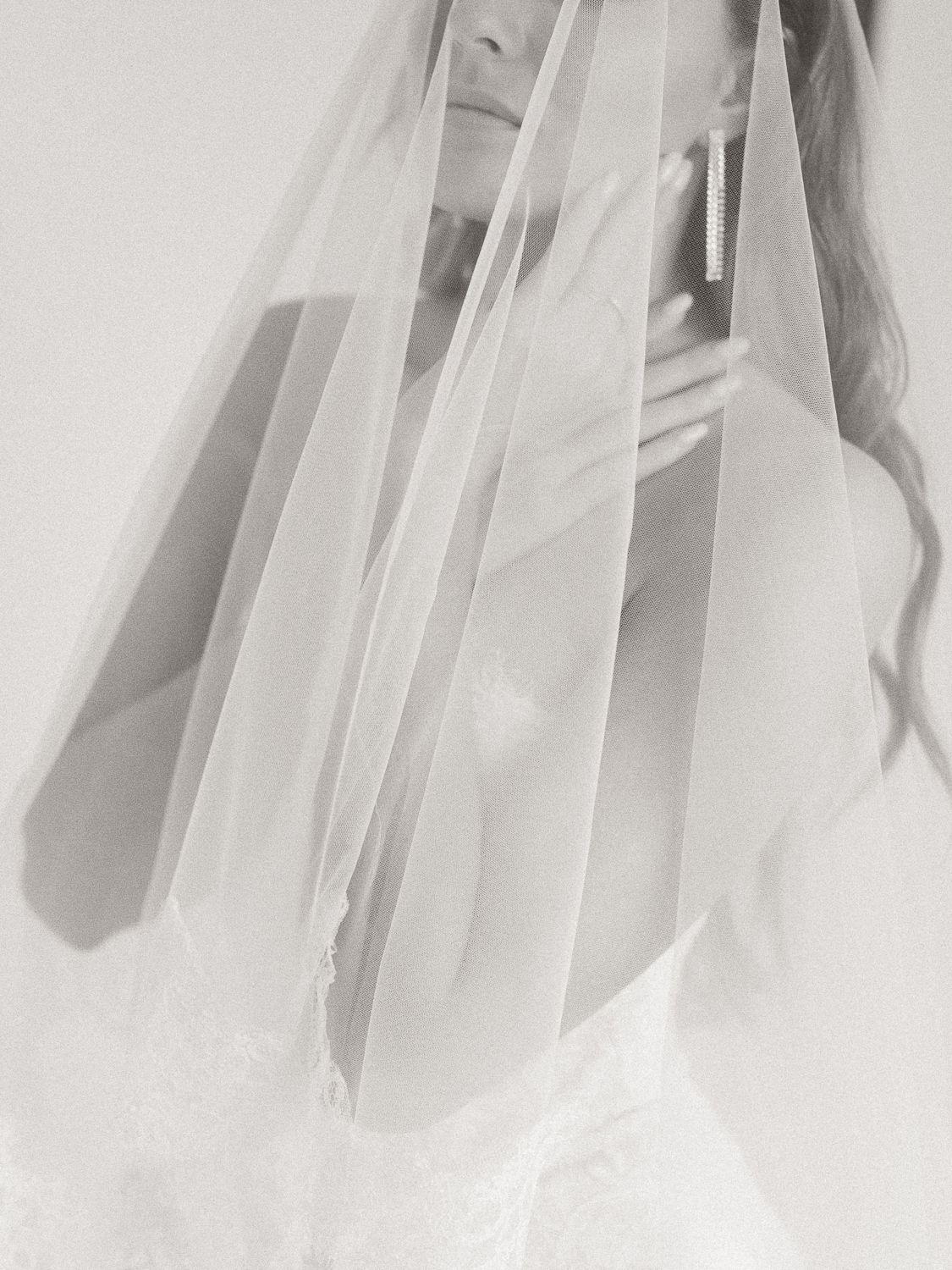 Black and white bridal portrait with flowing veil draped over bride wearing lace gown and elegant earrings.
