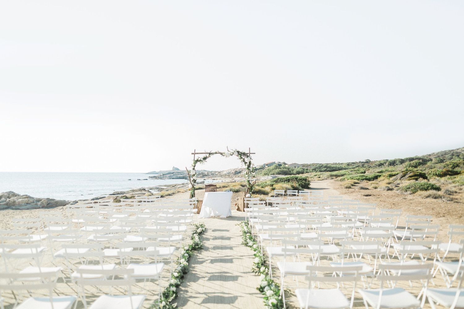 Elegant beachside wedding ceremony setup with white chairs, floral aisle, and rustic wooden arch overlooking the ocean.