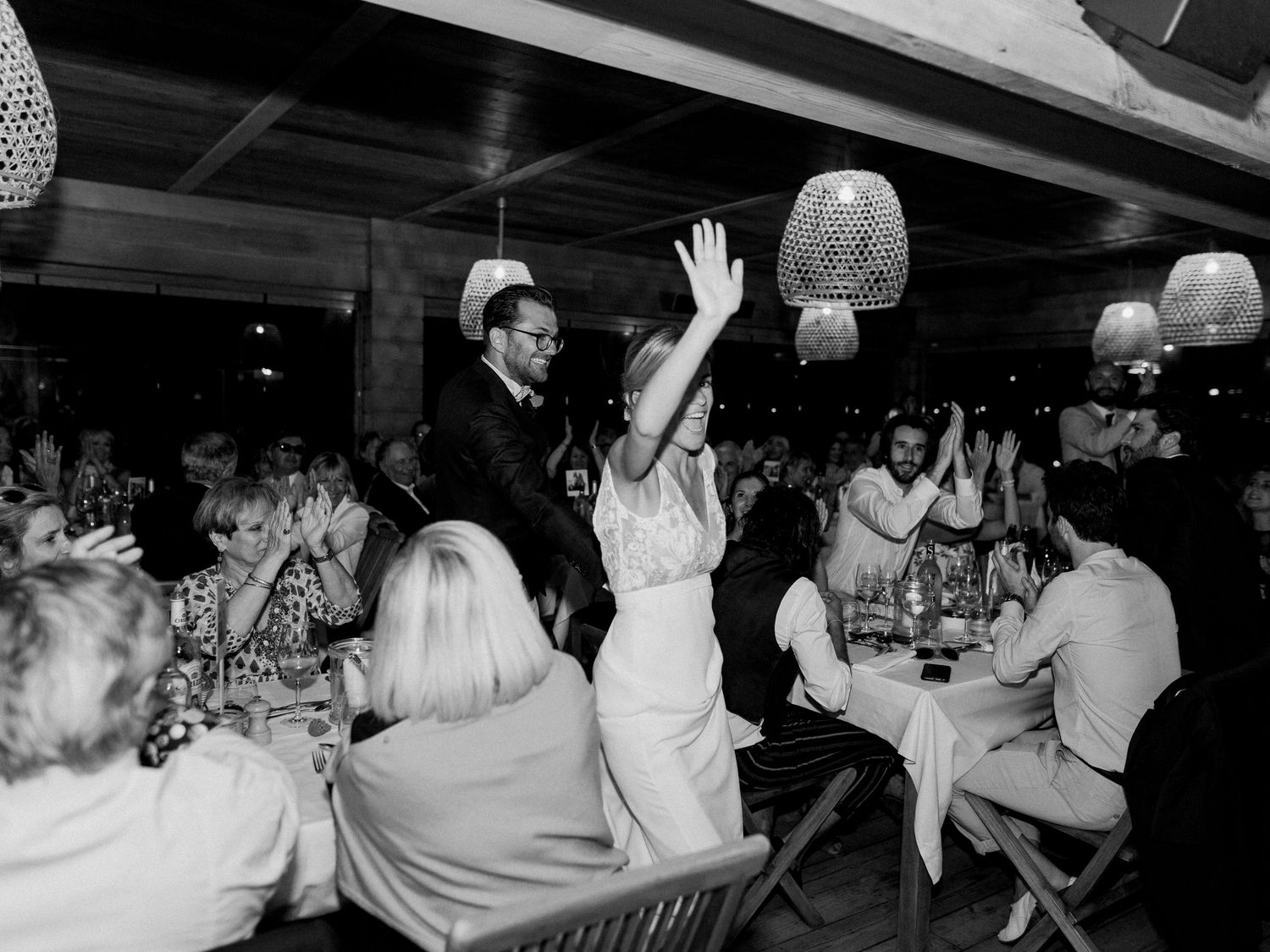Photo en noir et blanc d'un couple de jeunes mariés célébrant leur union, les bras levés, parmi leurs invités lors d'une réception festive