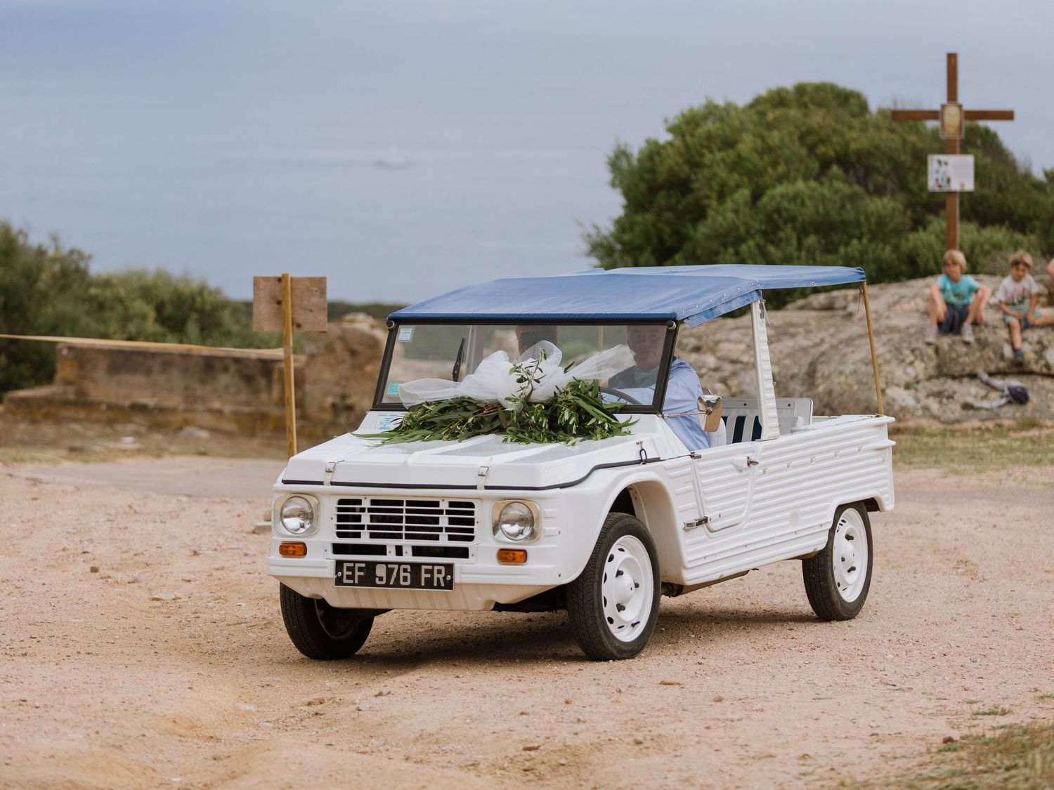 Une Citroën Méhari blanche, voiture de plage décorée de verdure, est garée sur un terrain sablonneux près d'une croix en bois