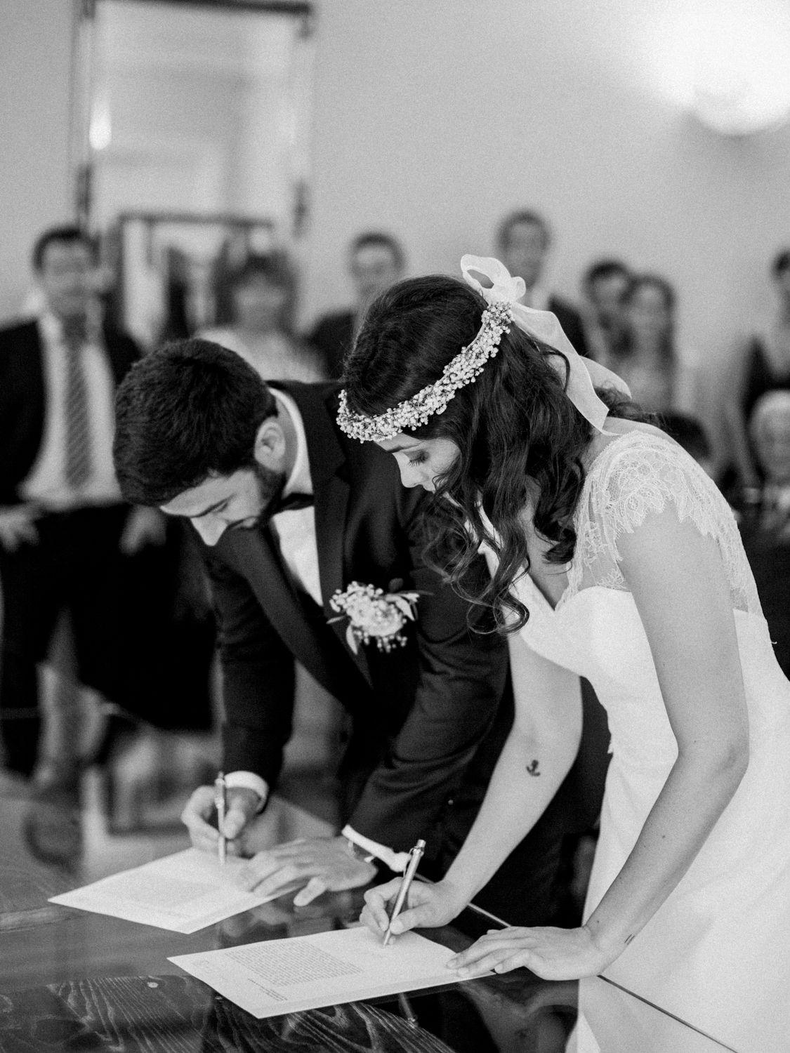 Bride and groom signing wedding certificate at ceremony table, guests watching in background, black and white photo.