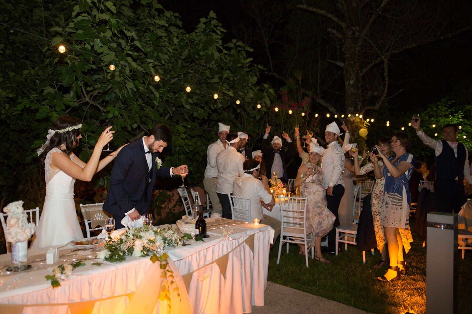 Bride and groom cutting wedding cake at elegant outdoor evening reception with string lights and cheering guests.