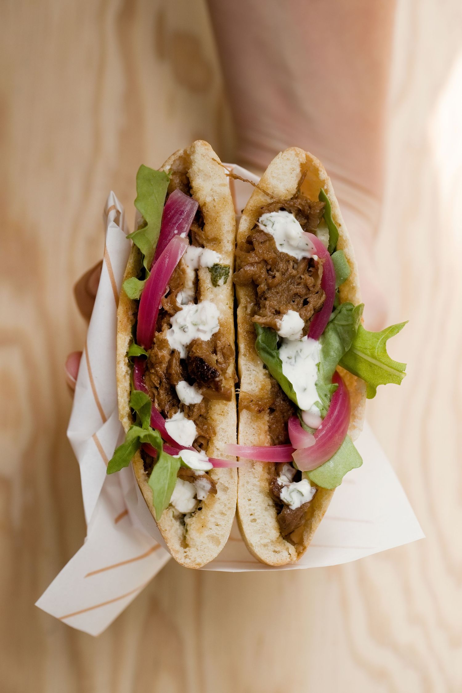 Des mains tenant un demi-sandwich pita gyro garni de viande, d'oignons rouges marinés, de légumes verts et de sauce blanche
à L'orriu.