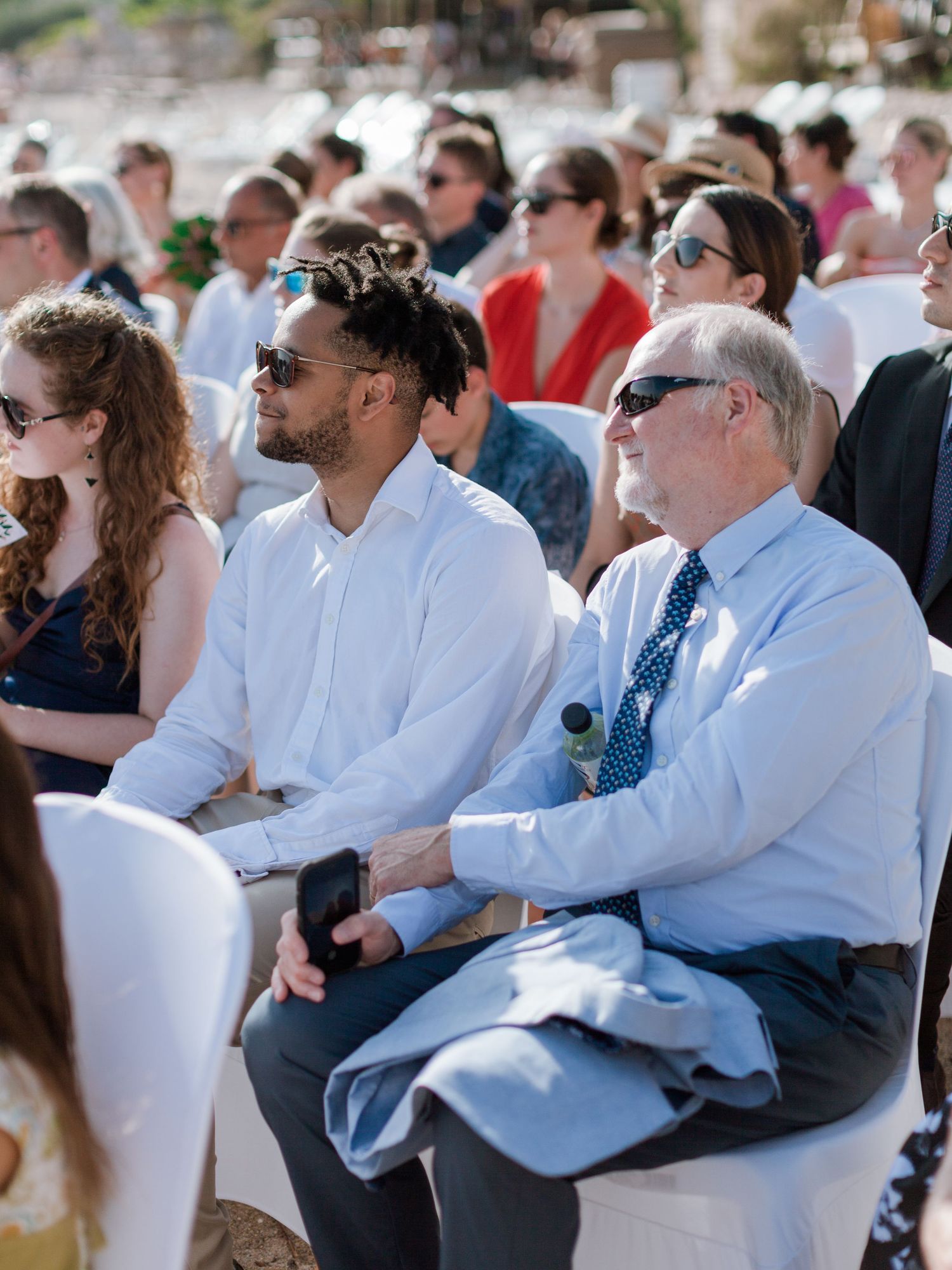 Invités assis en plein air lors d'une cérémonie de mariage ensoleillée sur la plage, portant des lunettes de soleil et une tenue habillée.