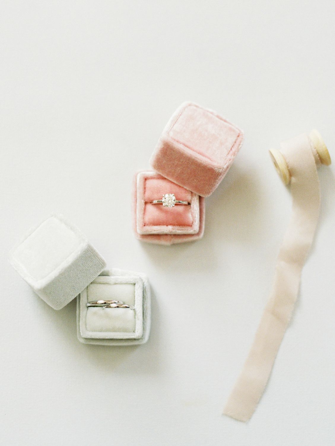 Two velvet ring boxes in pink and gray display elegant engagement rings beside a silk ribbon on a white surface.