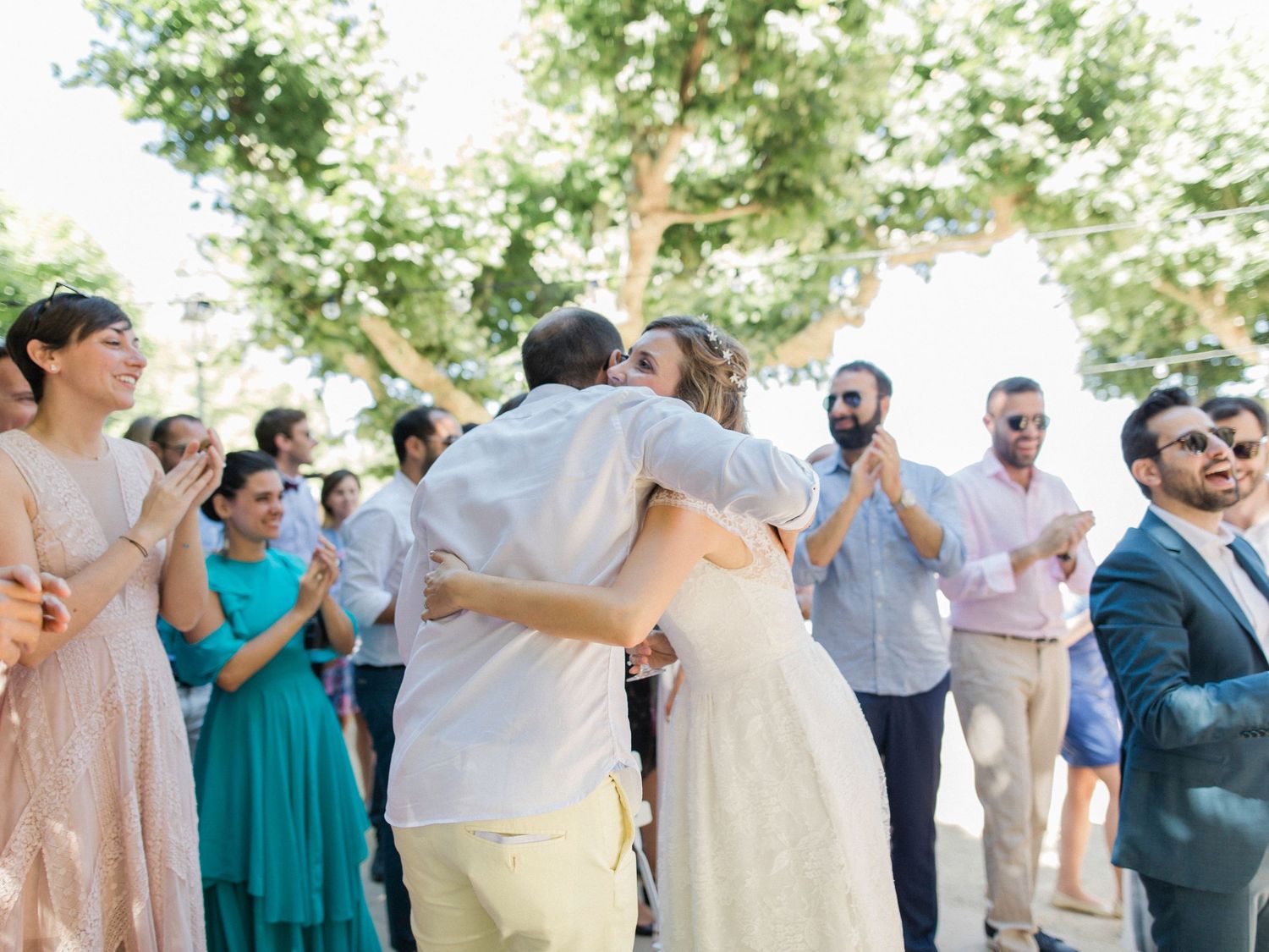 Couple shares first dance at outdoor wedding surrounded by clapping guests under green trees.